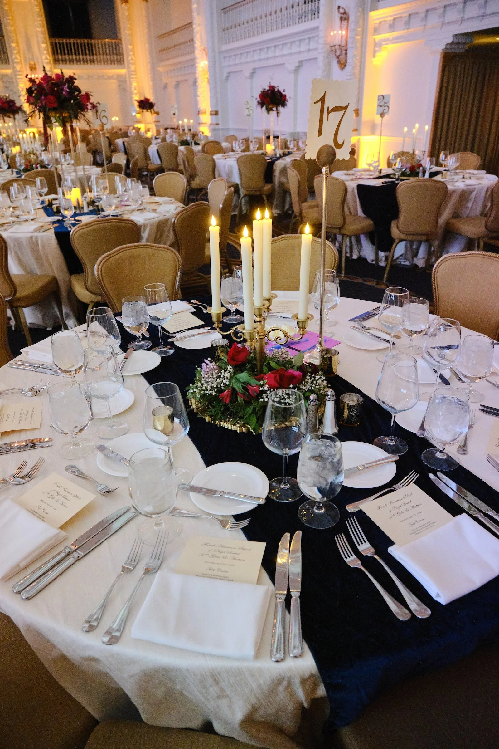 Elegant banquet hall set for a formal dinner with round tables decorated with candles and flowers, place settings, and menus, in a well-lit room with white walls and ornate details.