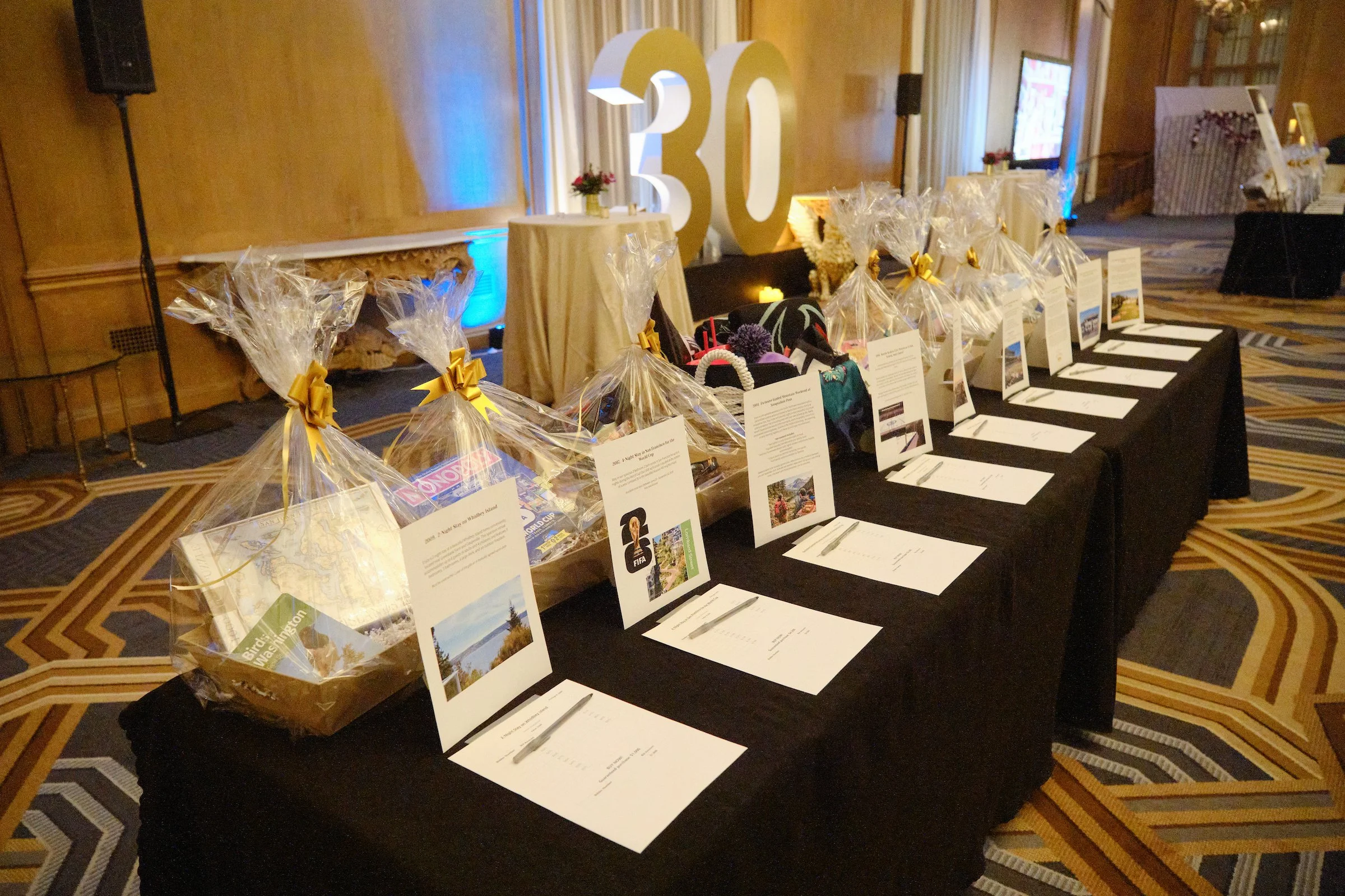 Event table with prizes wrapped in clear plastic with gold ribbons, certificates, and large golden '30' decoration in the background at a celebration event.