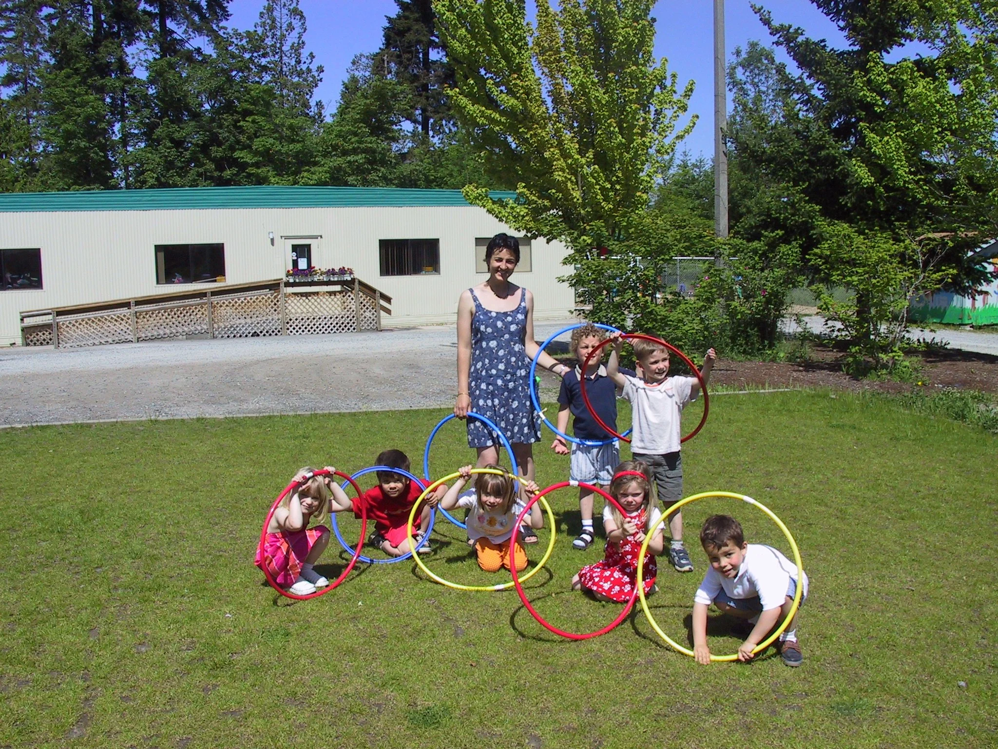 A group of young children and three adult women smiling in a playground with wood structures and greenery in the background.