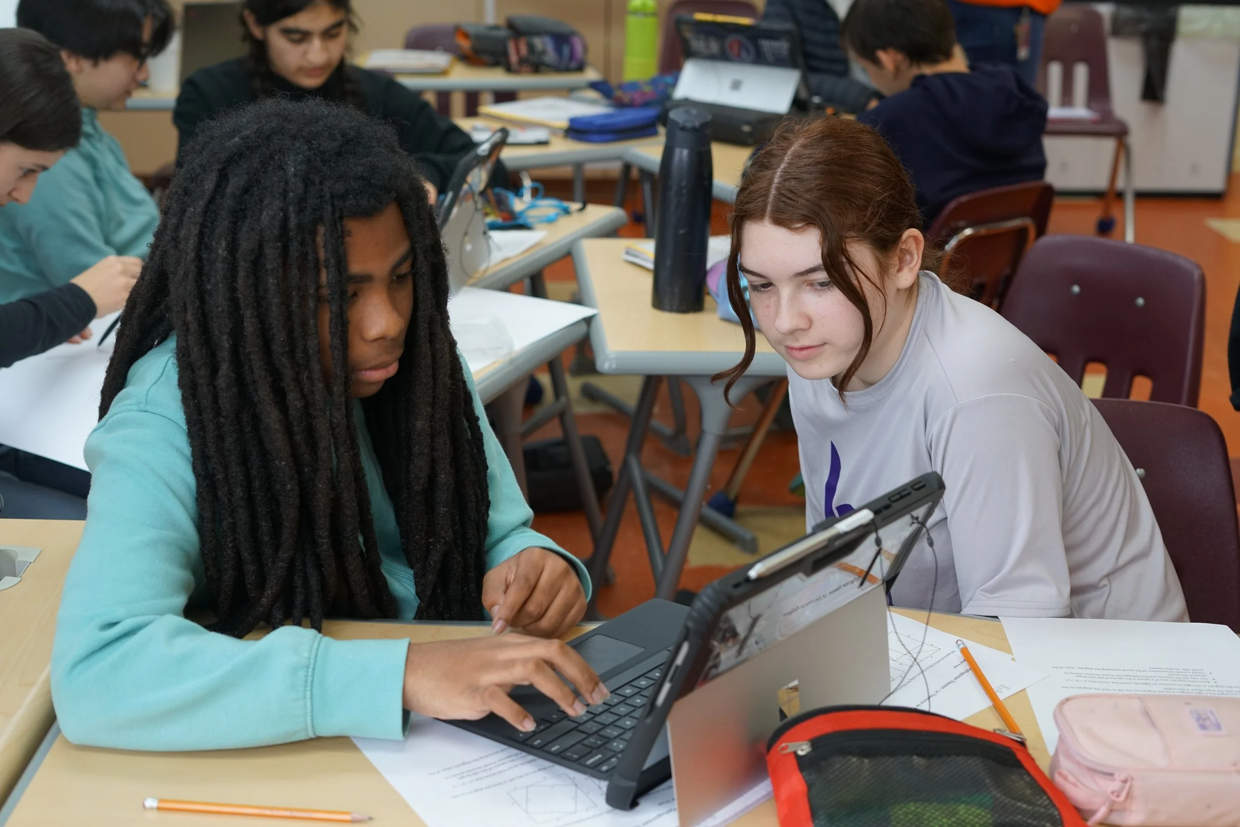 Two students, a girl with long dreadlocks and a girl with red hair, are working together on a laptop in a classroom. Other students are visible in the background.