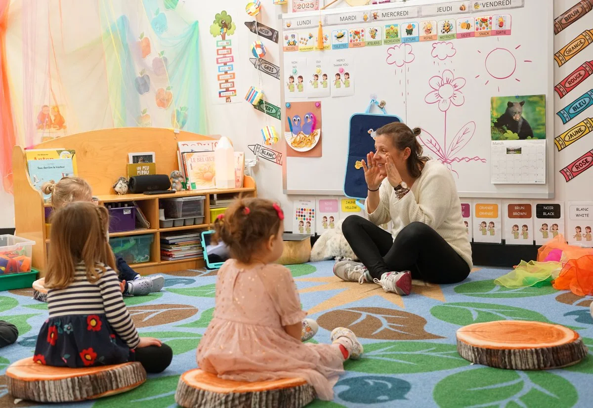 A teacher sitting on the floor with children in a classroom, engaging them in a lesson. The children are sitting on small wooden stools, with colorful educational decorations and whiteboard drawings in the background.