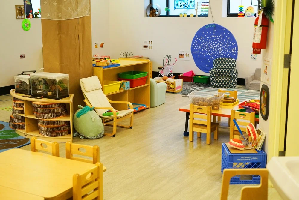 A colorful classroom with small wooden tables and chairs, storage bins, a white chair with a plush pillow, and an area for crafts and activities with toys and decorations.