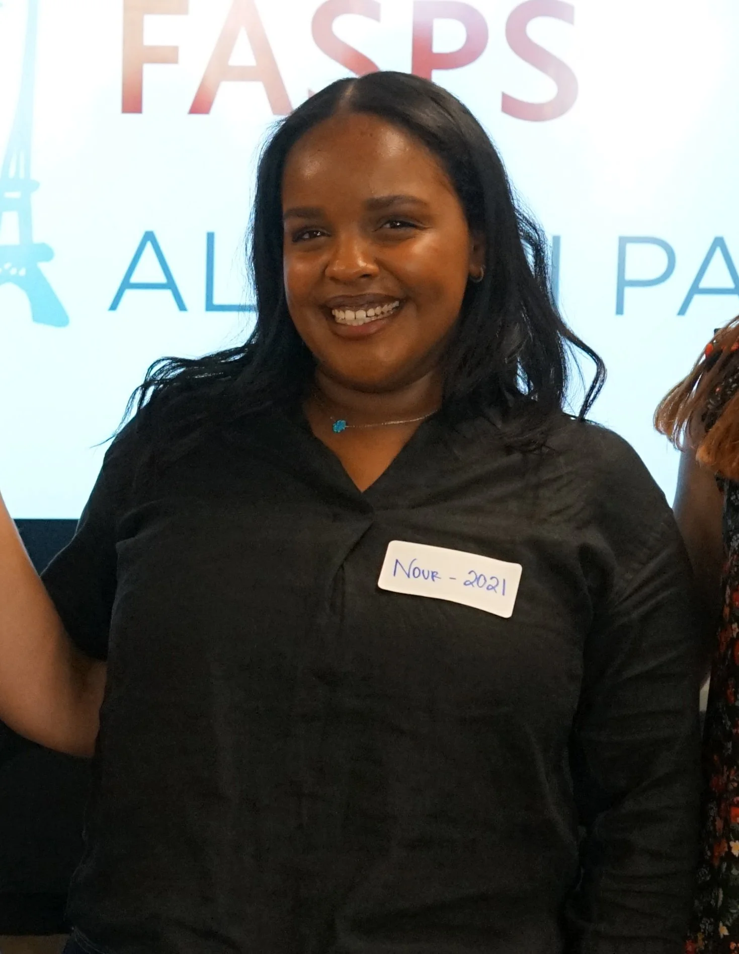 A smiling woman wearing a black top and a blue necklace, standing in front of a sign that says 'FASPS.' She has a white name tag that says 'NOUR - 2021.'