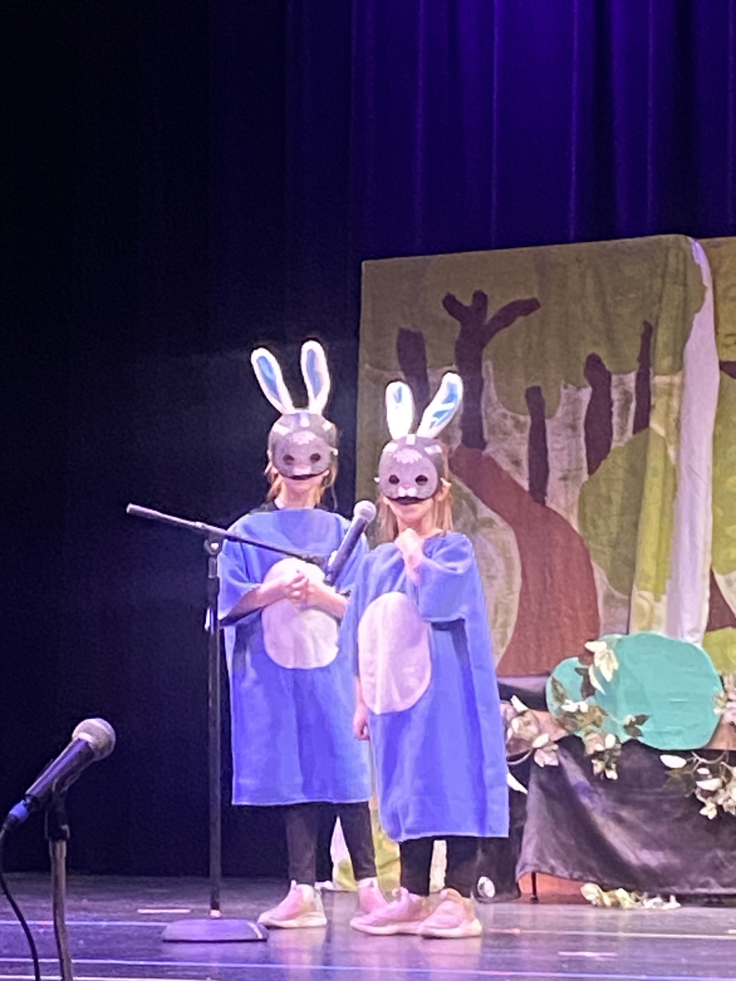 People wearing bunny masks, blue costumes, and bunny feet on stage with a painted backdrop and flowers on the floor, performing in a play or show.