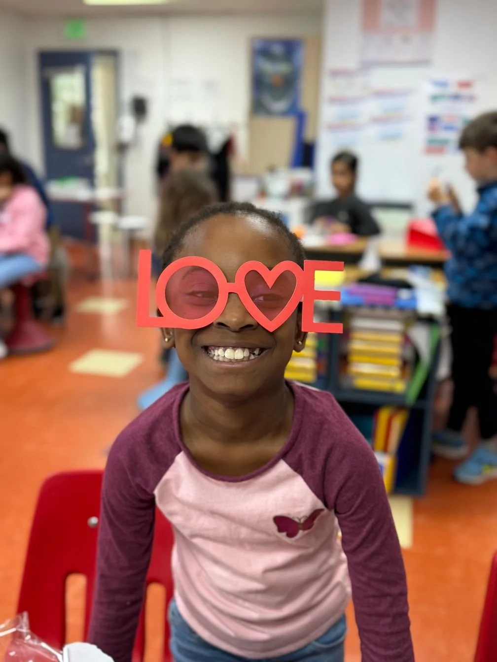Smiling girl in a classroom holding a red 'LOVE' sign over her face with the 'O' replaced by a heart shape.