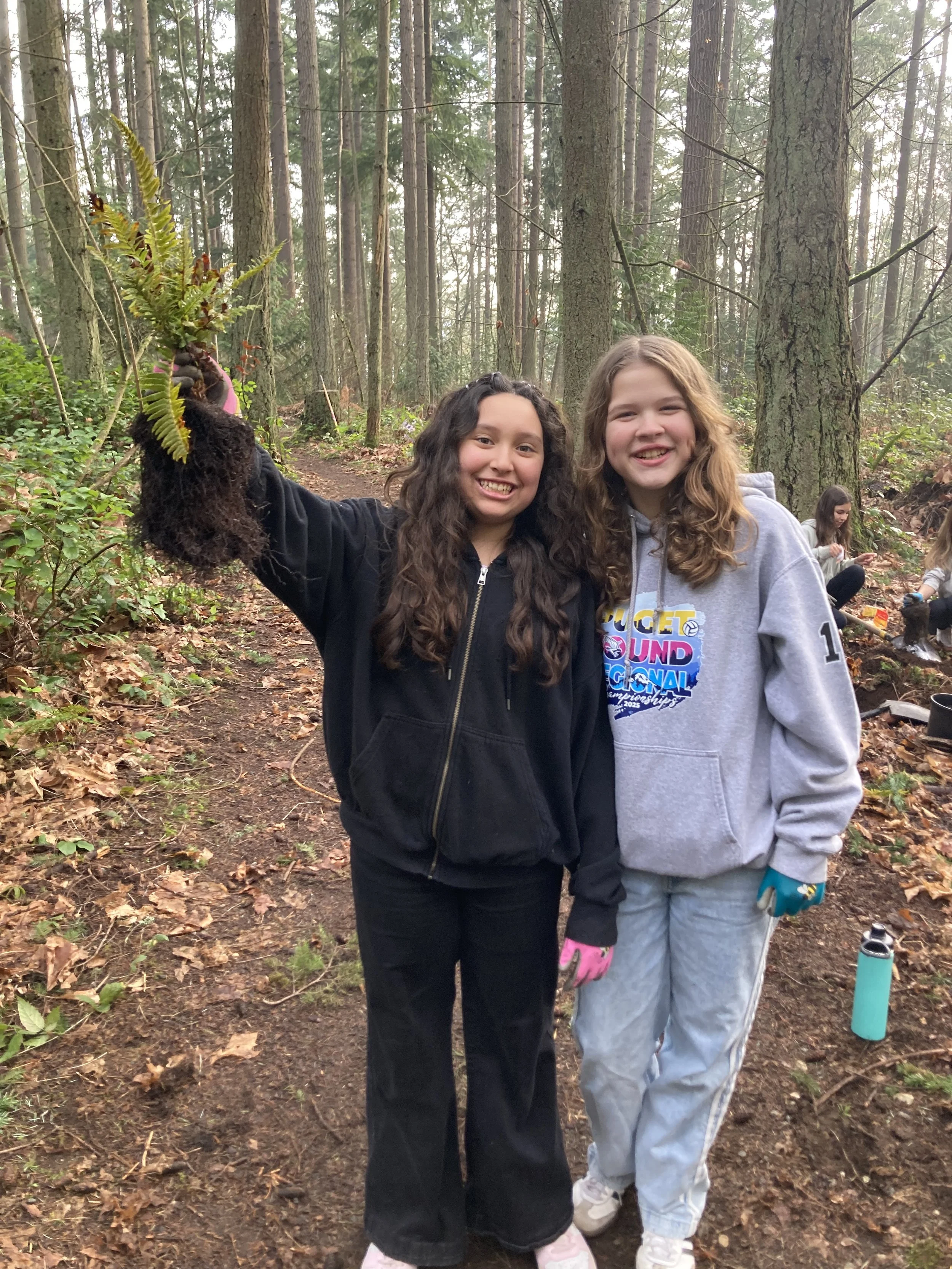 Two girls smiling and standing in a forest, one holding a fern plant. Other children are seen in the background, sitting on the ground with plants and water bottles around.