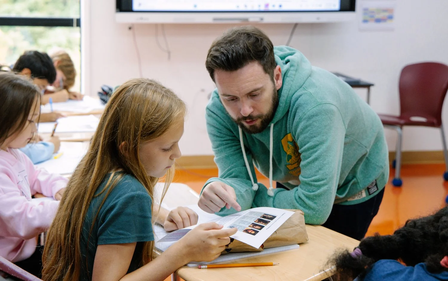 A teacher helping a young girl with reading or homework in a classroom with other students.