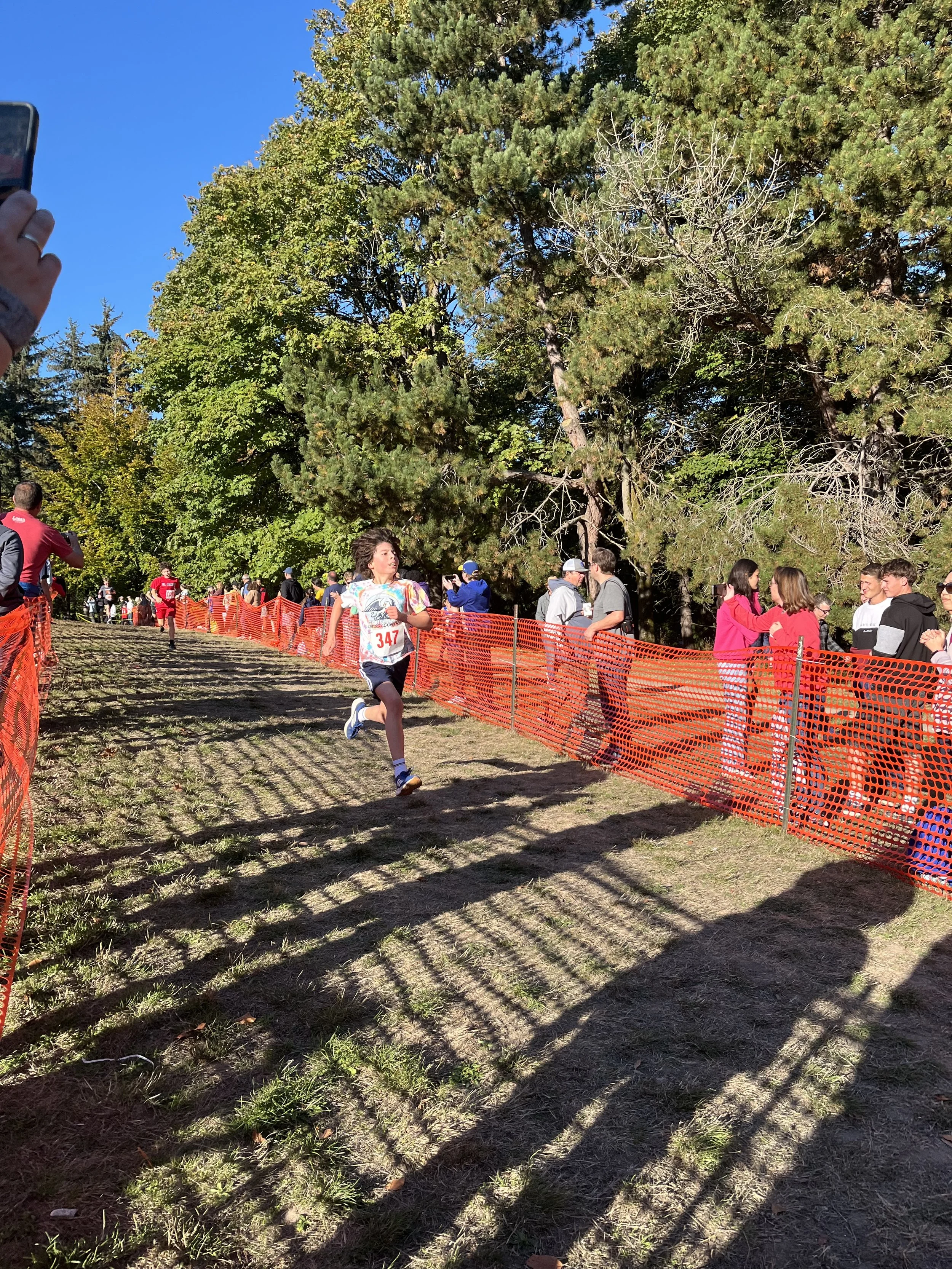 A group of people participating in a running event along a trail lined with orange safety fencing, surrounded by tall trees on a sunny day.