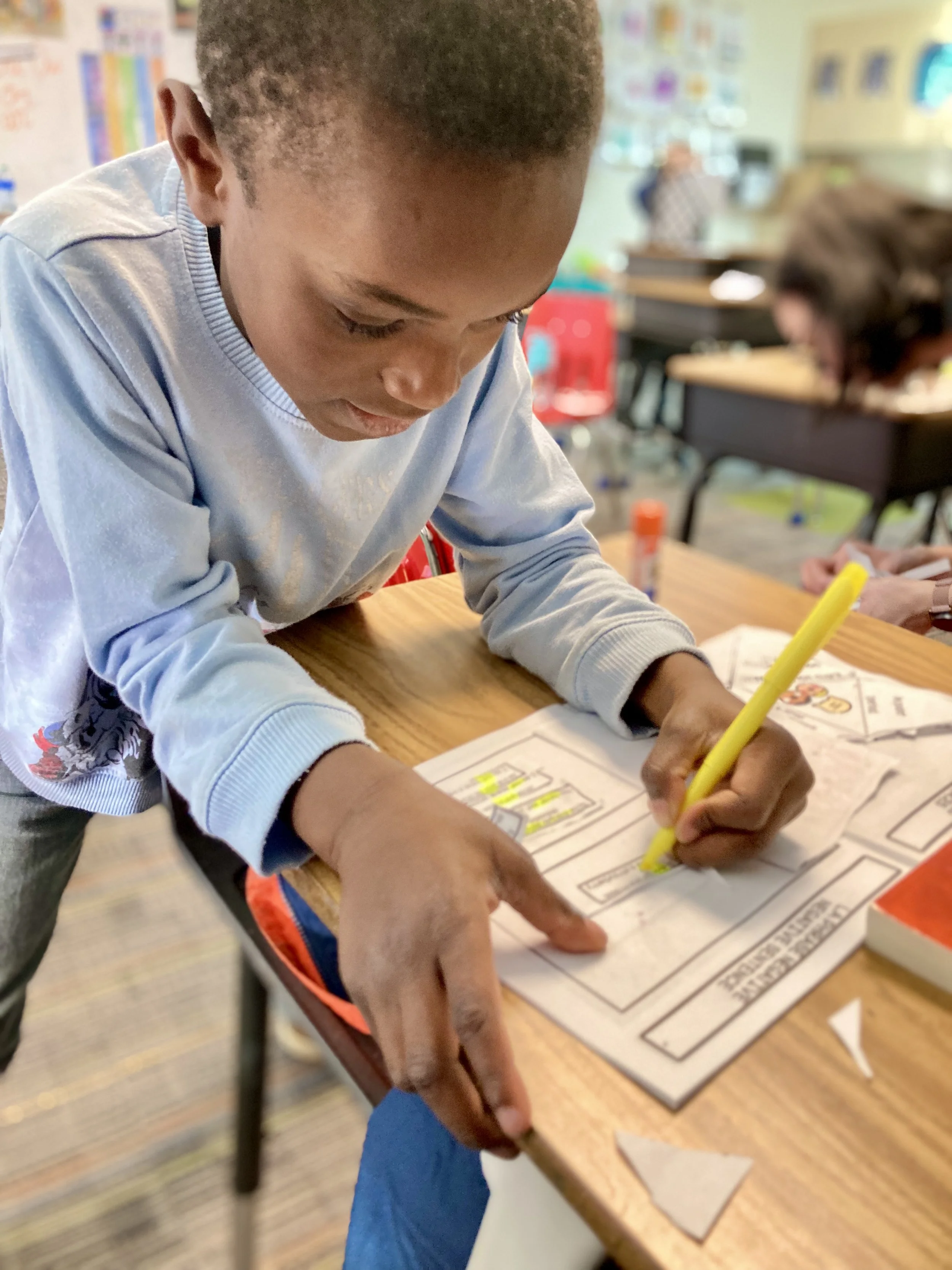 A young boy with dark hair and a light gray long sleeve shirt writing with a yellow marker on a worksheet on a wooden desk in a classroom. There are other children and classroom materials in the background.