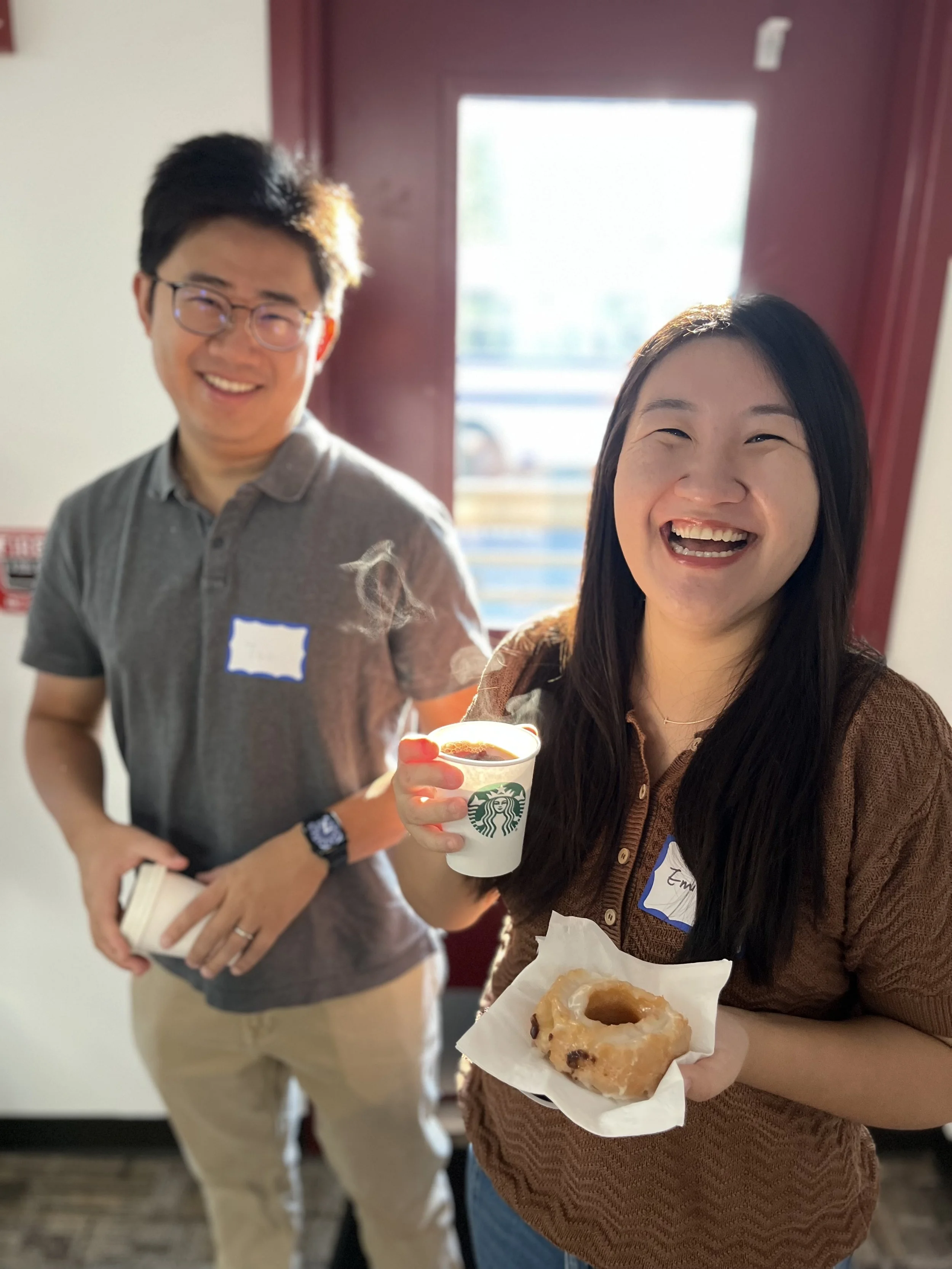 Two people smiling, one woman holding a donut and a coffee cup, a man standing in the background with a book, inside a room with a window letting in natural light.