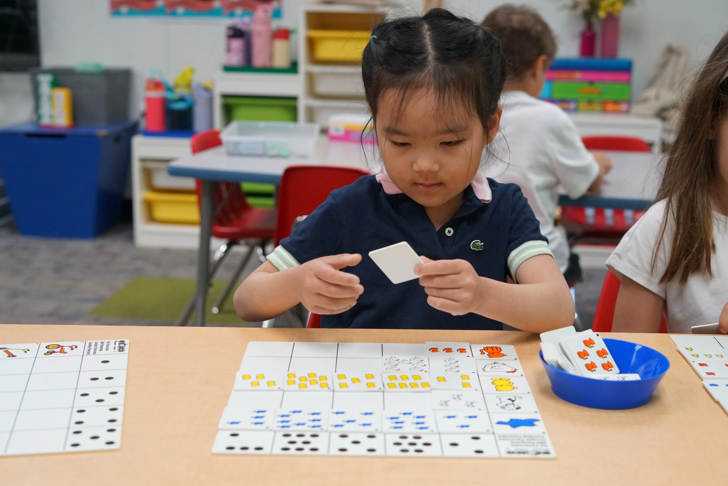 A young girl sitting at a classroom table looking at a white card. On the table are educational materials including number cards, dice, and colorful counters. Other children and classroom shelves with books and supplies are visible in the background.
