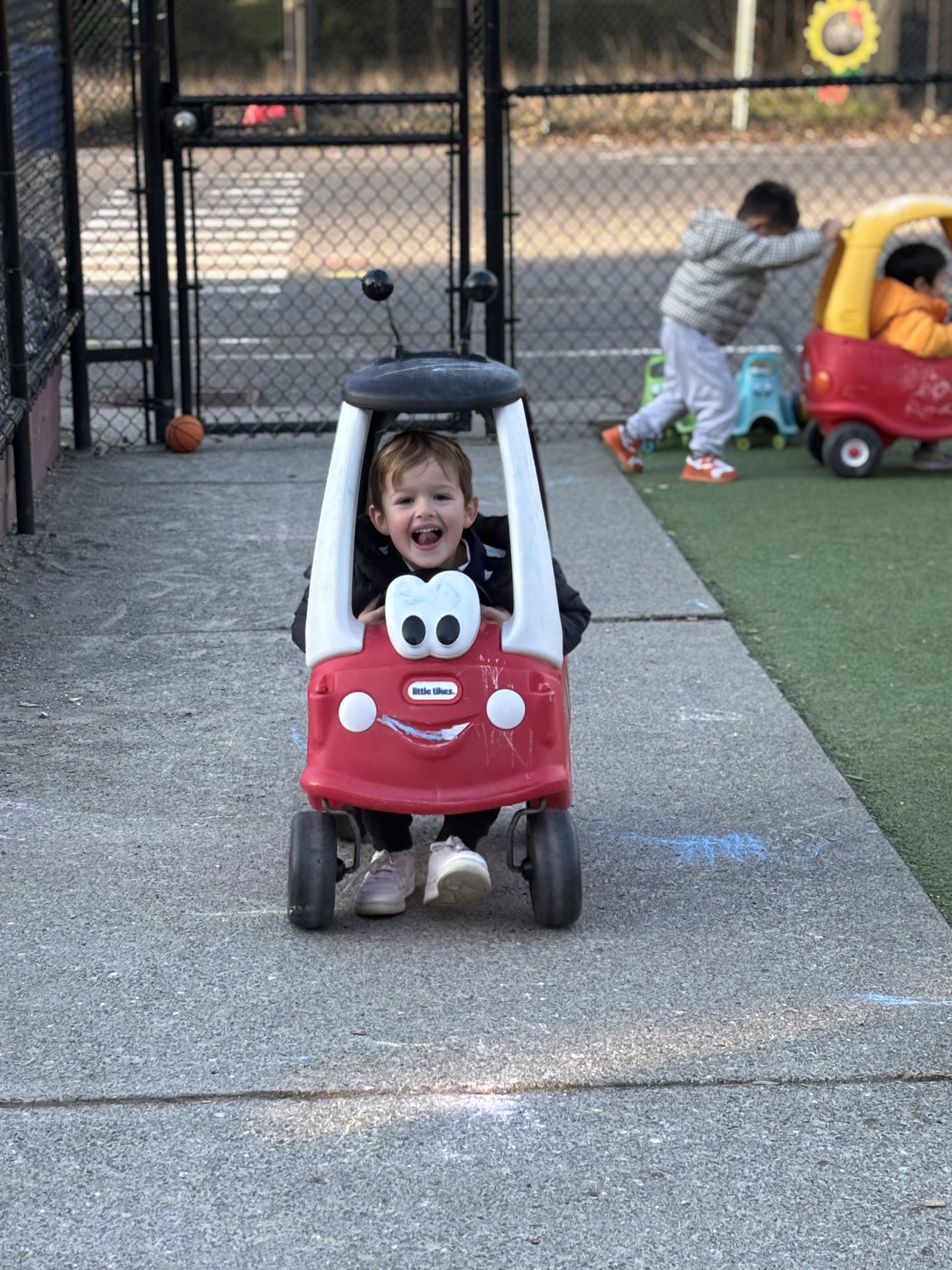 A young child with brown hair smiling and sitting in a red toy car shaped like a character with white eyes and a smiling face, at a playground. In the background, other children are playing near a fence, with one child leaning against a yellow and red plastic play structure and another in a blue and green toy vehicle.