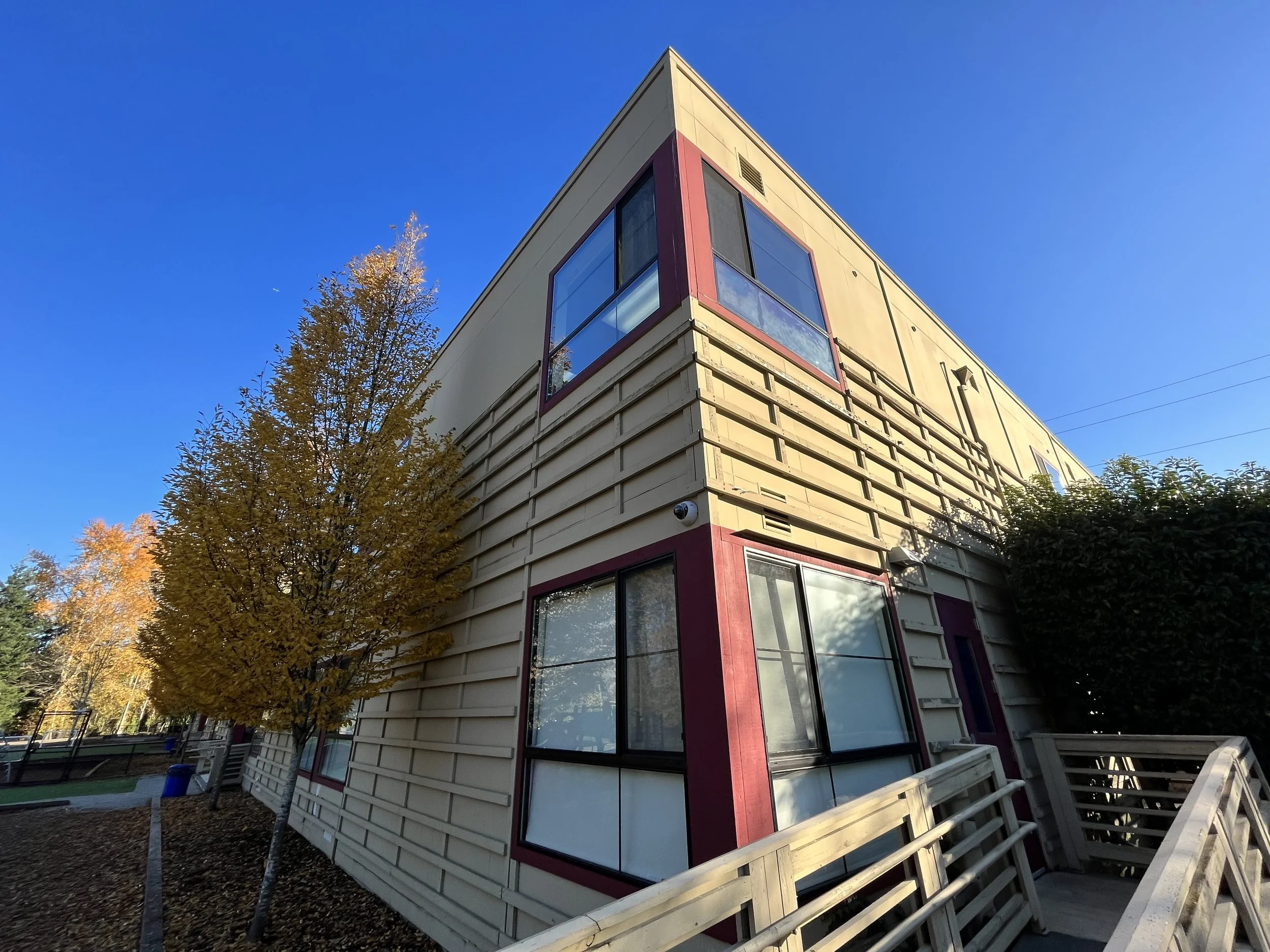 A multi-story building with beige siding and red window frames, surrounded by trees with autumn foliage, set against a clear blue sky.