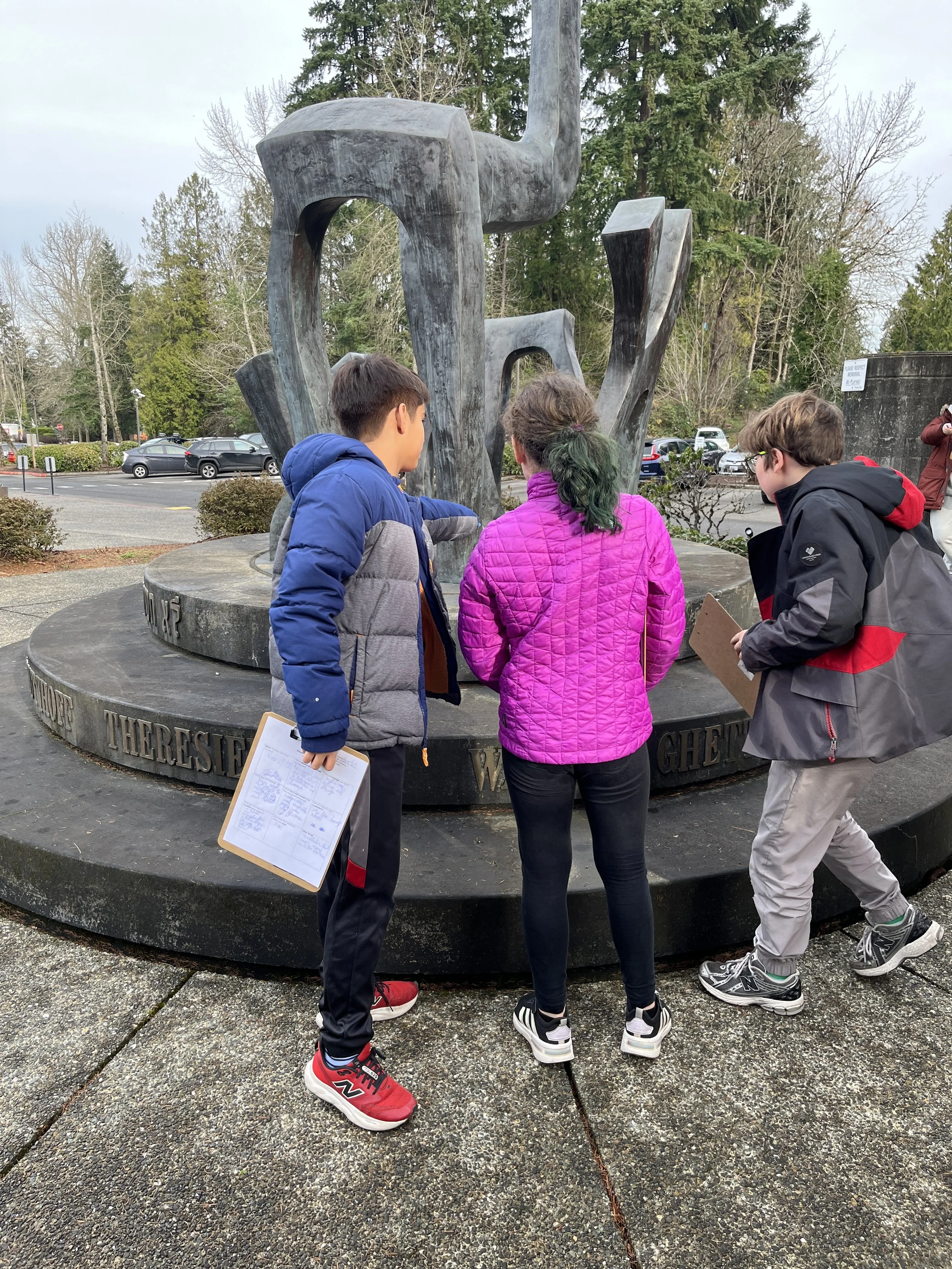 Three children standing around a large outdoor monument with metal sculptures and inscriptions, with a parking lot and trees in the background.