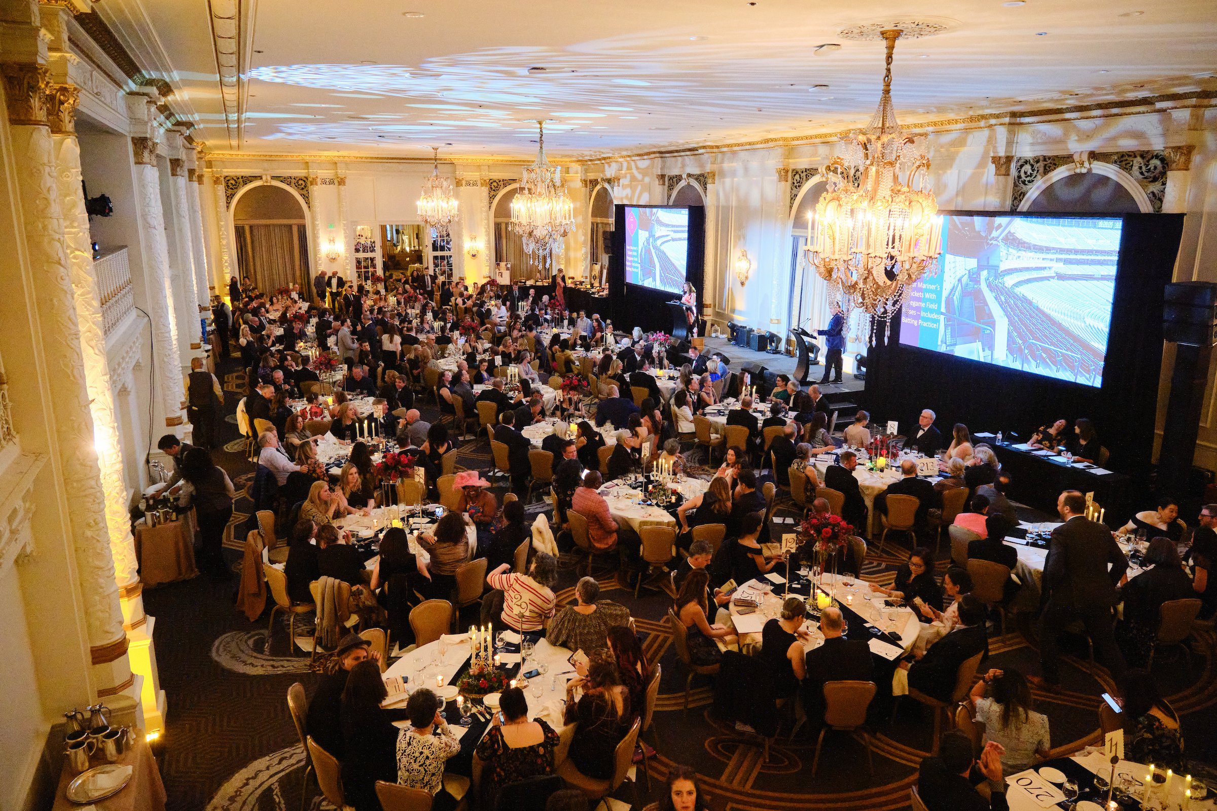 An elegant banquet hall filled with many round tables, each set with fine dinnerware and floral centerpieces, hosting numerous formally dressed guests attending an event or celebration. Large screens and a stage with a speaker are visible at the fron