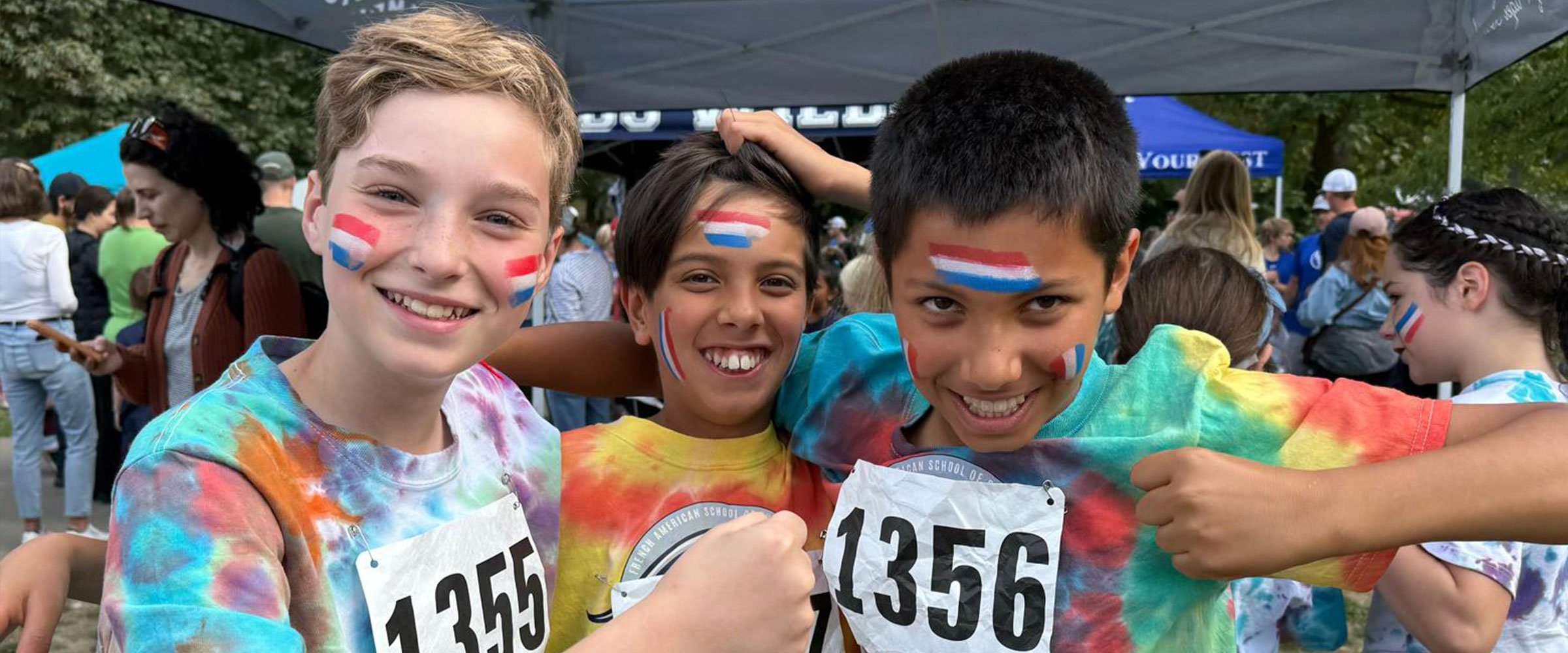 Three young boys with face paint and colorful tie-dye shirts at a race event, smiling and showing medals outside under a tent, with other people in the background.