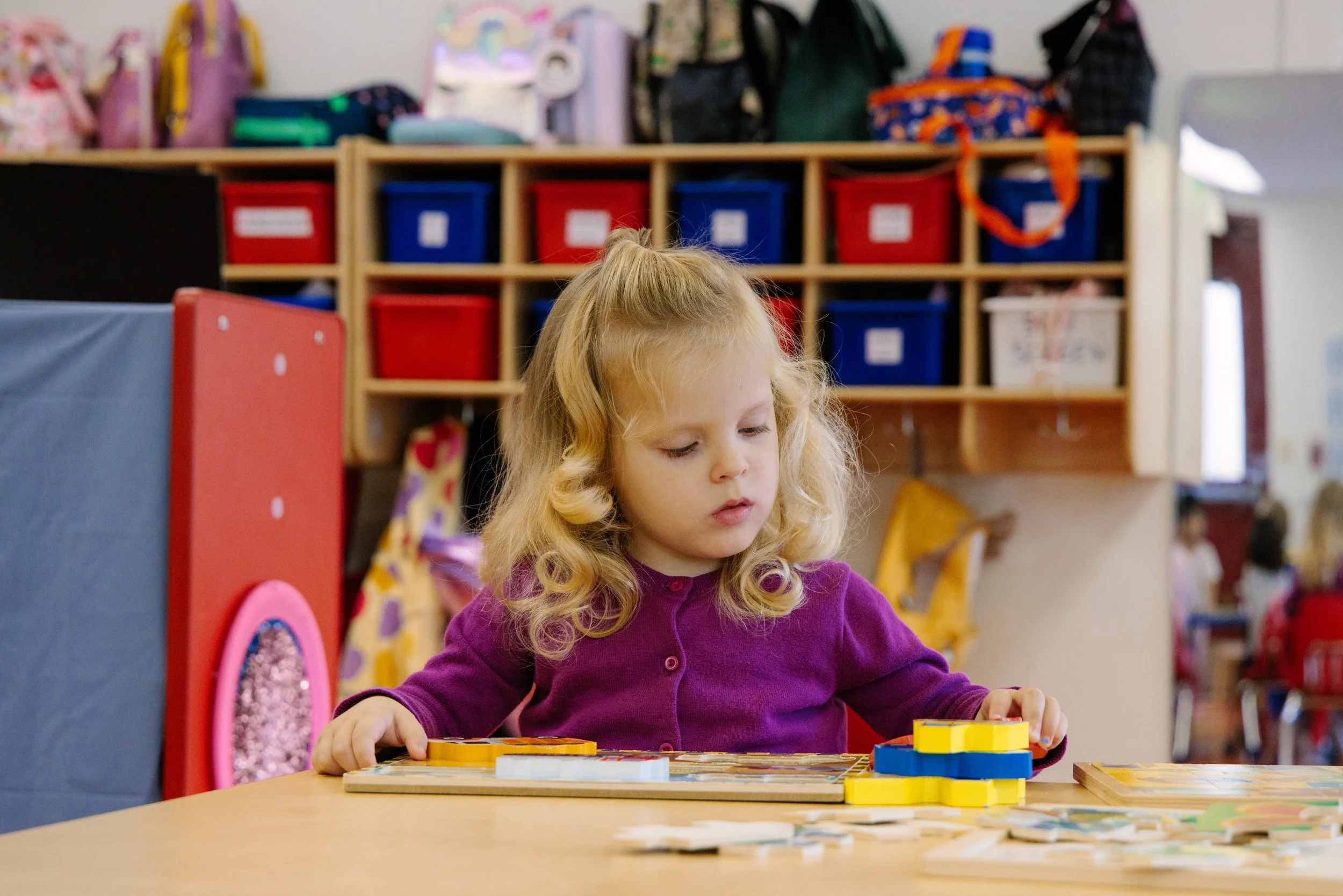 A young girl with curly blonde hair wearing a purple sweater playing a game at a table in a classroom or playroom.