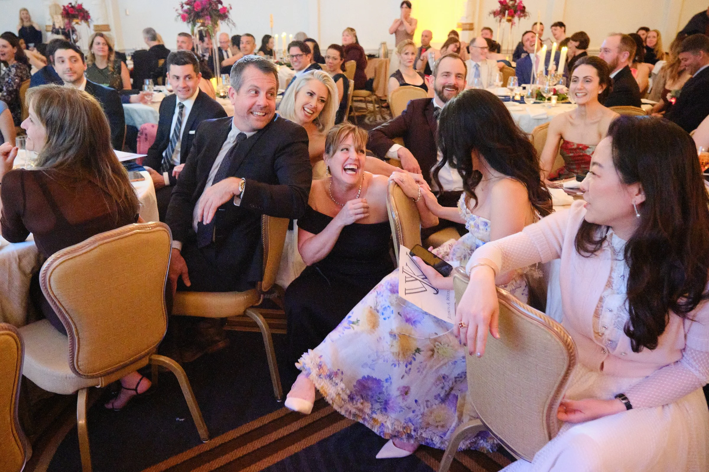 Group of people at a formal event, smiling and laughing, seated around banquet tables with fancy floral centerpieces and candles in a well-lit room.