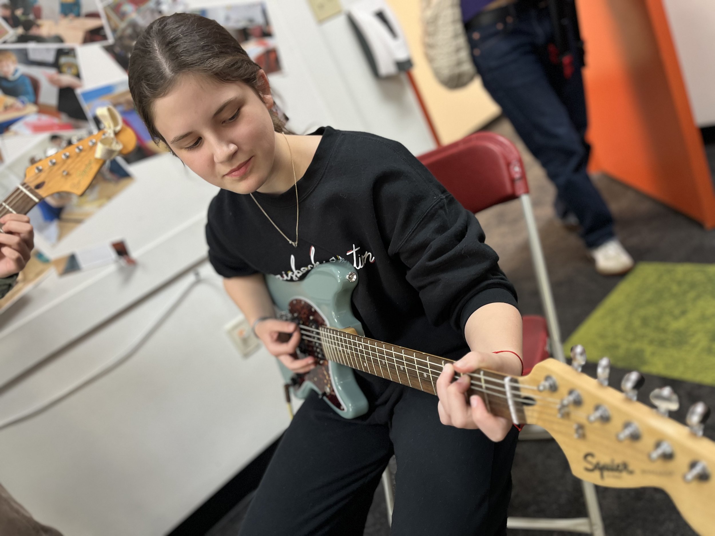 A young person with short brown hair playing an electric guitar in a music classroom, with posters and other students in the background.