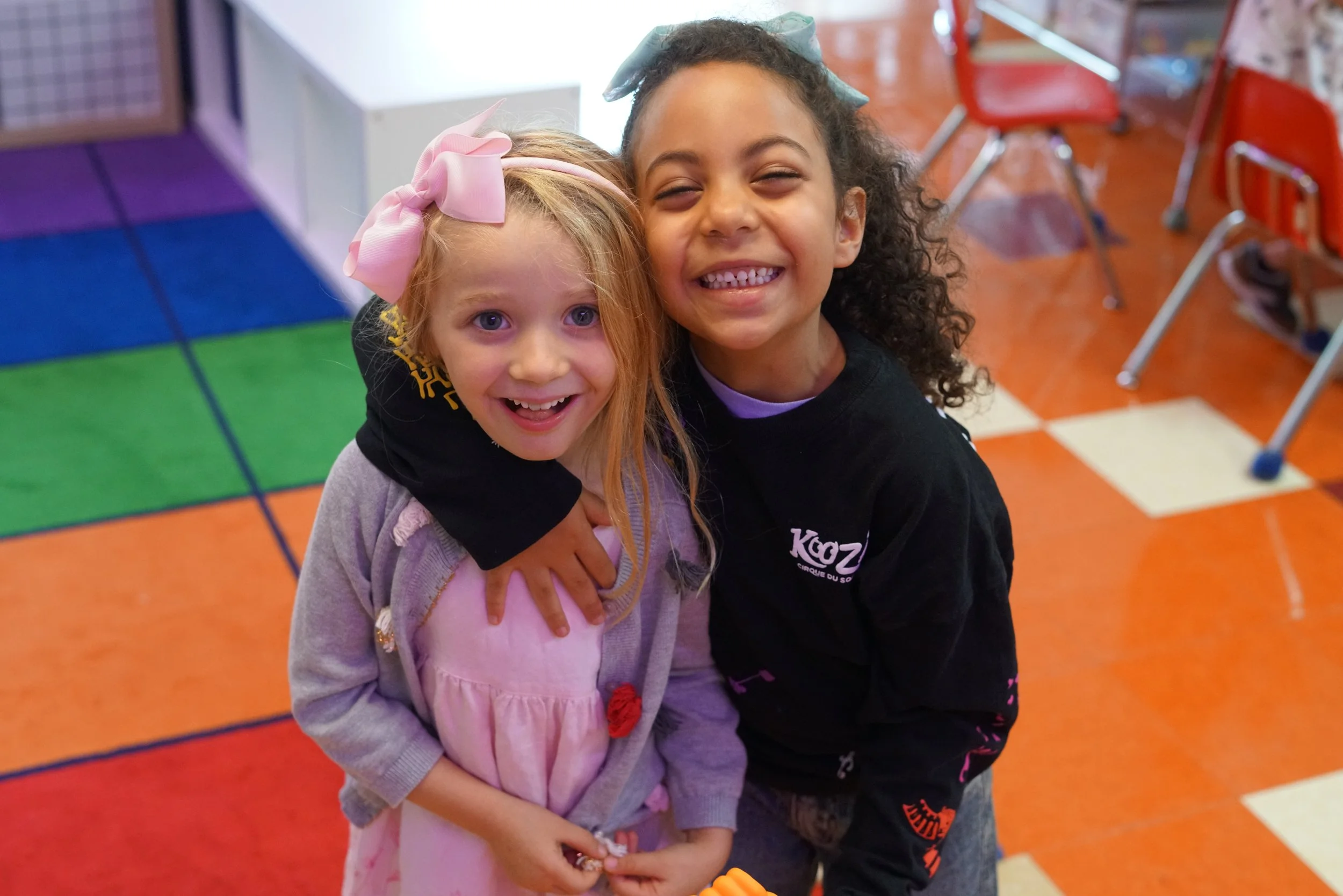 Two young girls hugging and smiling inside a colorful indoor play area with orange floor tiles, red chairs, and a multicolored mat.