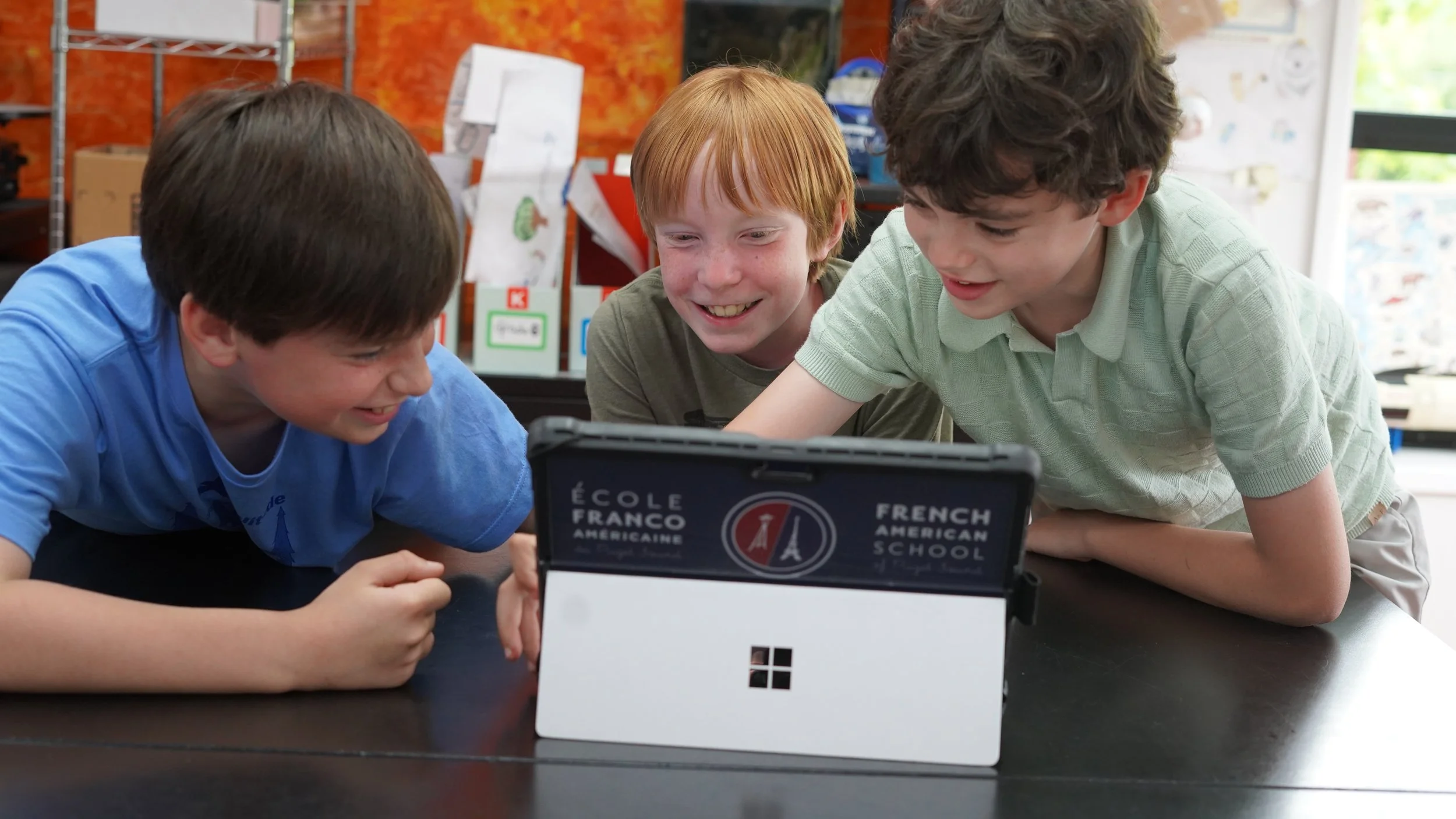 Three boys are gathered around a small tablet or device on a table, smiling and looking at the screen. One boy with brown hair is on the left, another boy with red hair is in the middle, and a third boy with curly brown hair is on the right. The device has a label that reads 'Ecole Franco Americain' and 'French American School.' The background shows some shelves, papers, and a window with natural light.