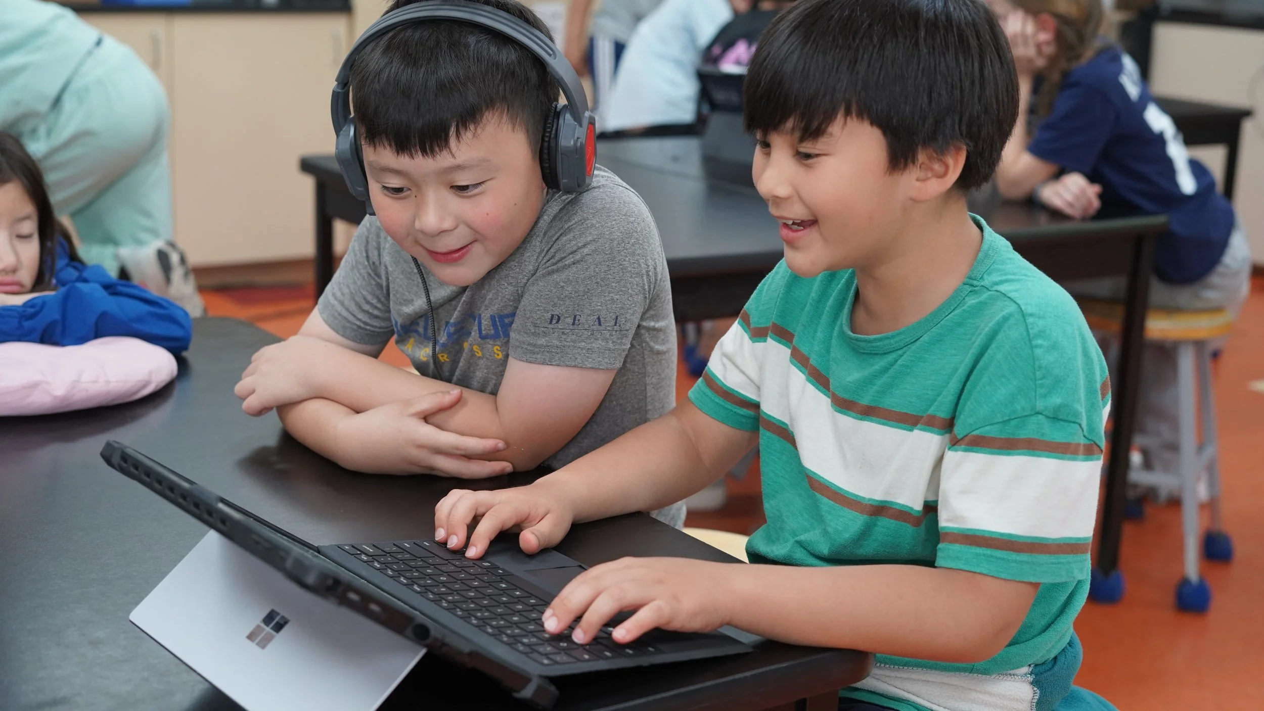 Two boys working together on a laptop in a classroom, with other students in the background.
