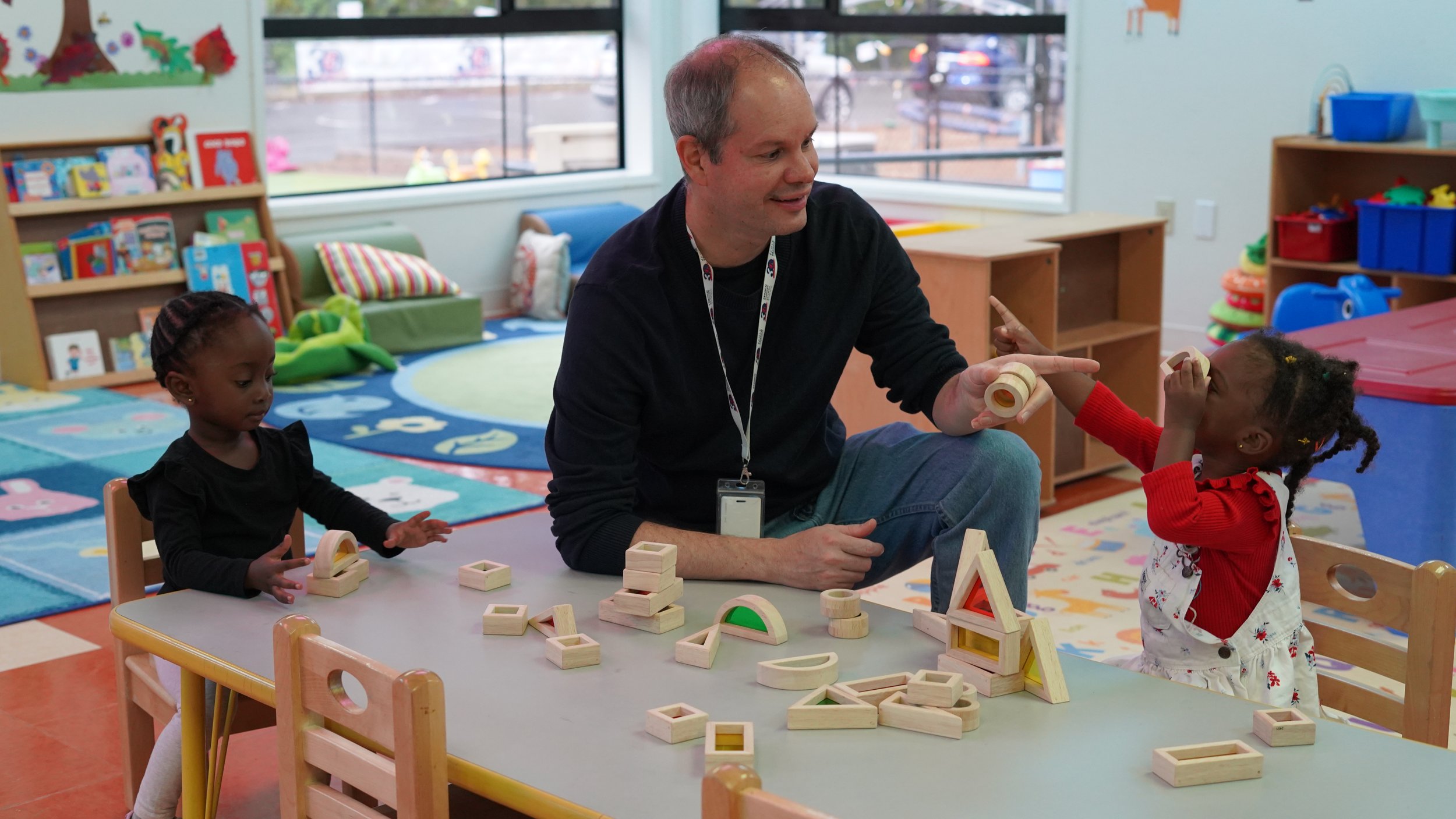 An adult man and two young girls playing with wooden building blocks at a table in a colorful classroom.