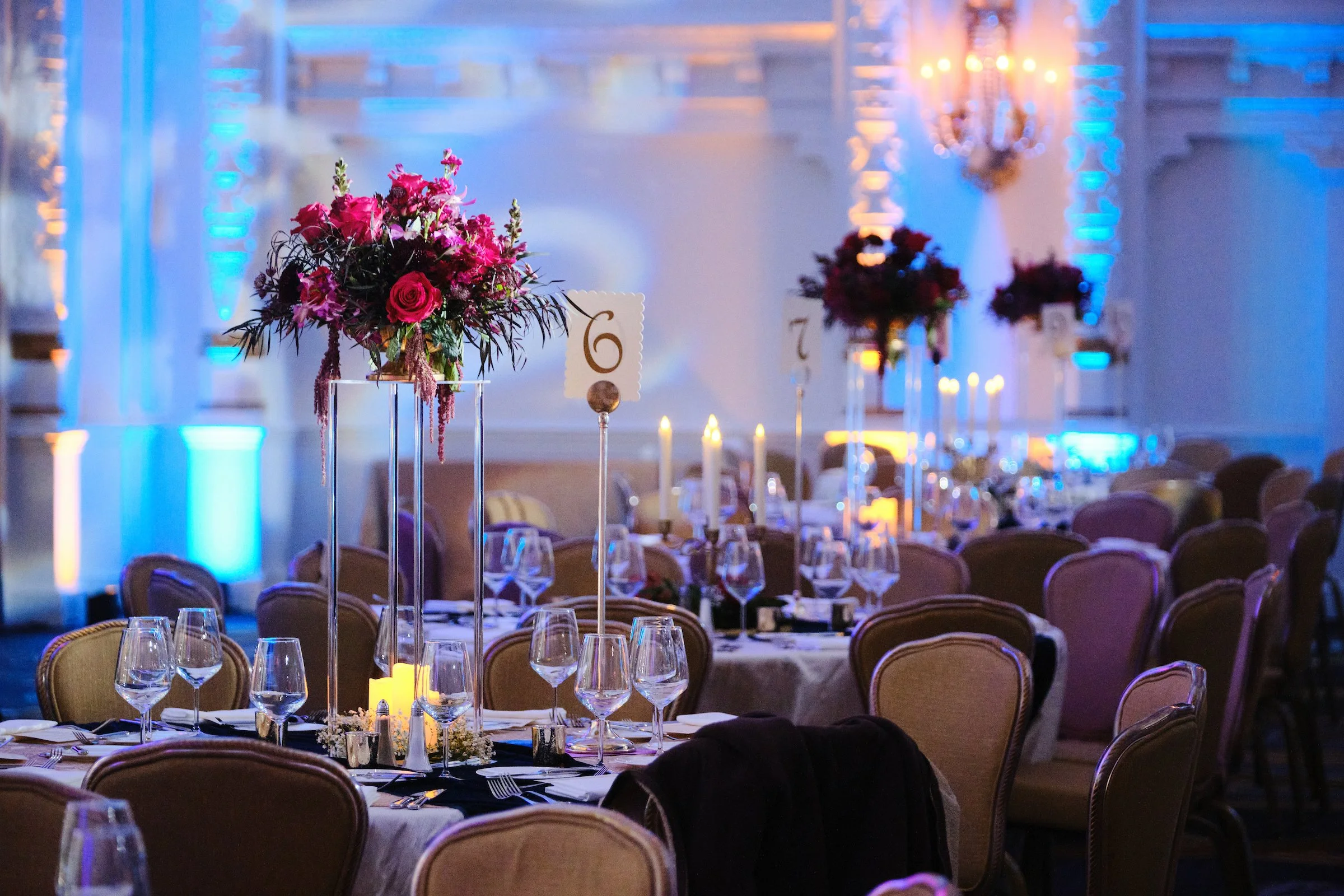 Elegant banquet hall decorated with large floral centerpieces featuring pink and red flowers on high stands. The room is set with wine glasses, plates, and silverware on round tables covered with white tablecloths. The background has blue and purple 