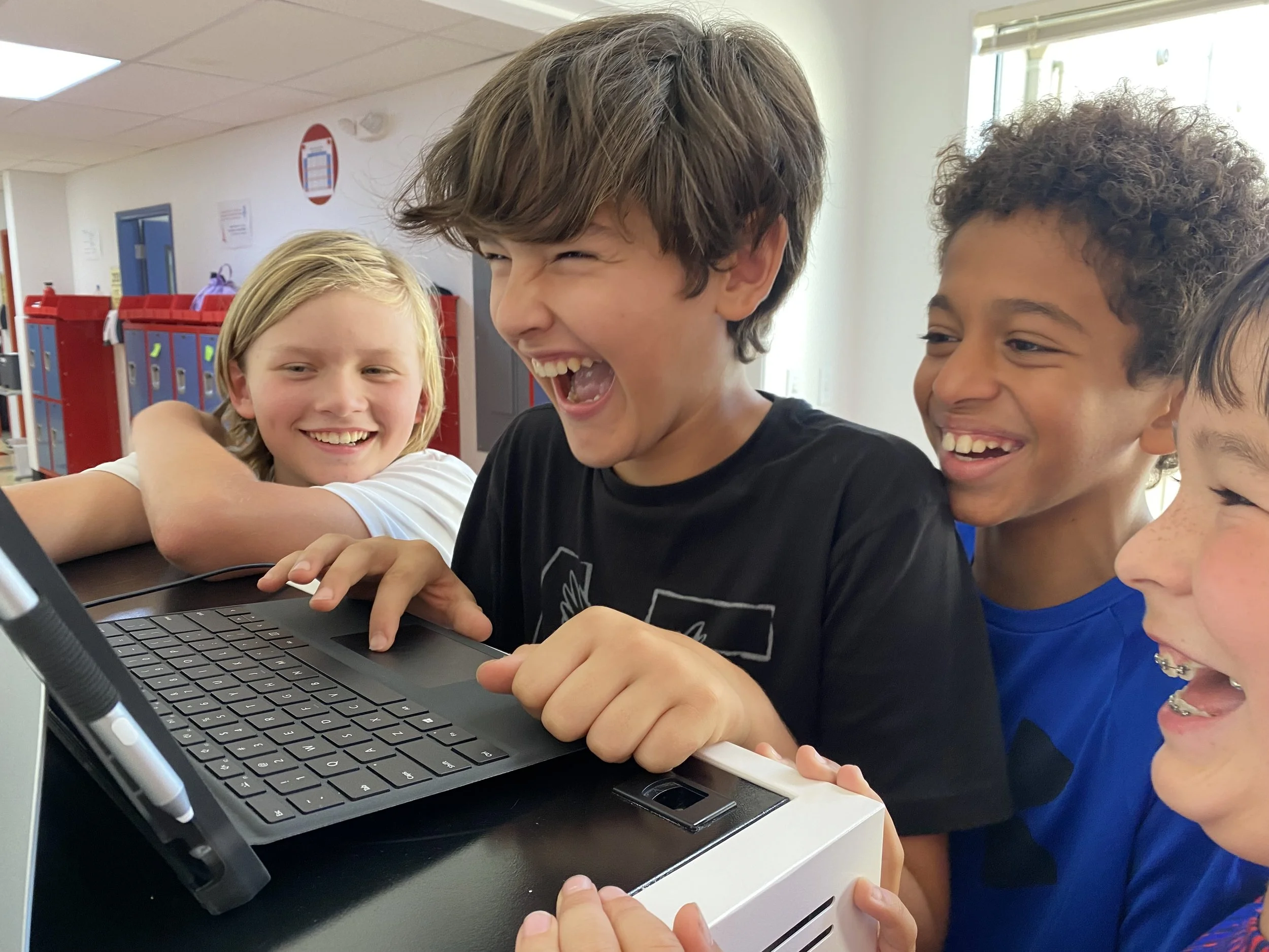 A group of children gathered around a laptop computer, laughing and smiling together, in a room with lockers and educational posters.