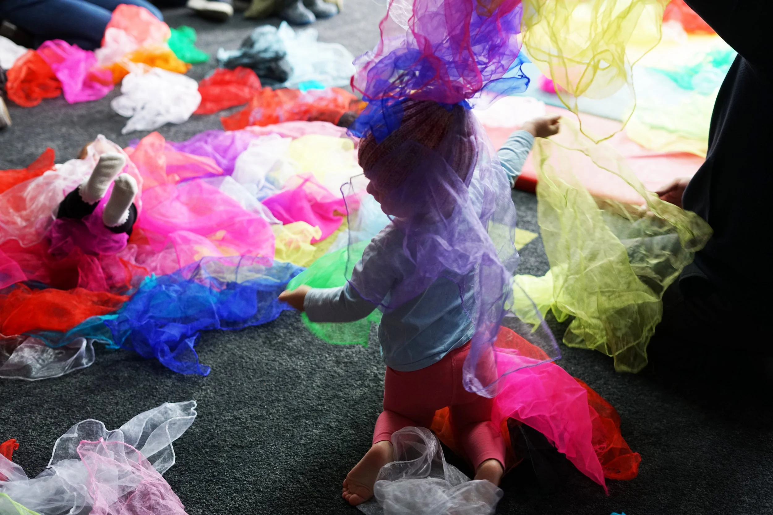 Children playing on a floor with colorful netting and fabric pieces, surrounded by scattered fabric and netting in various bright colors.