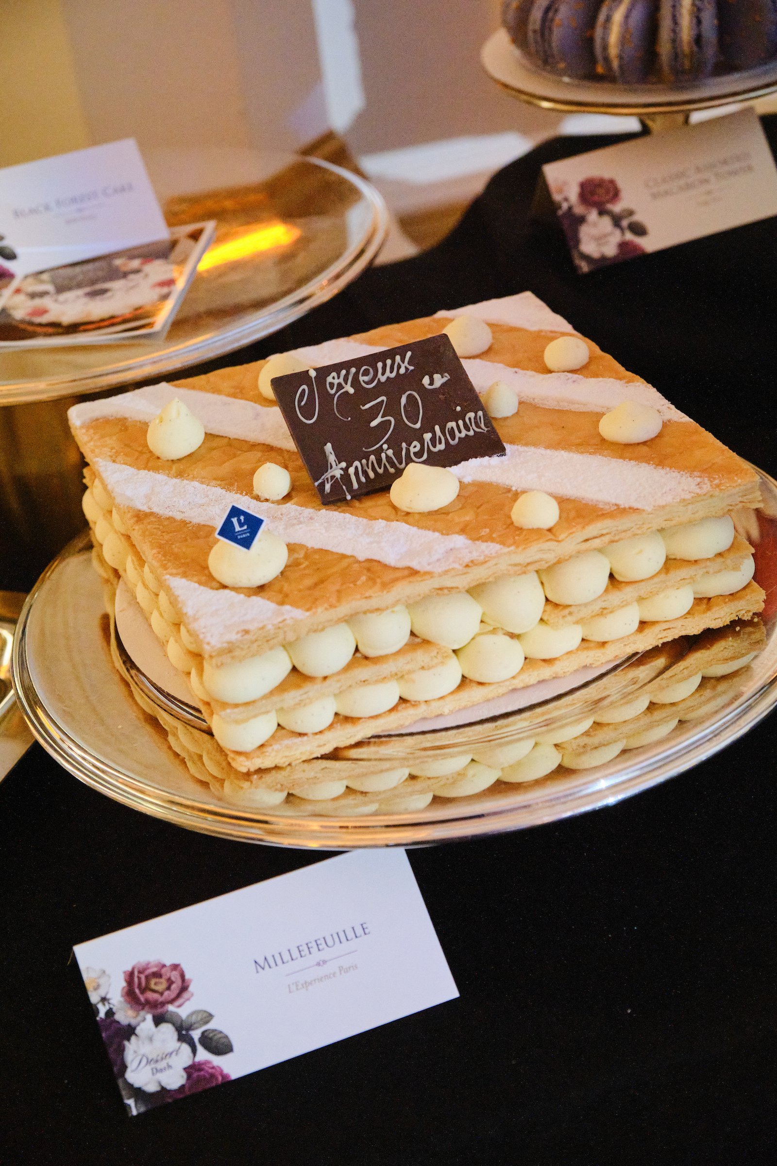 A rectangular millefeuille cake decorated with white cream dots and powdered sugar, with a small chocolate sign reading '30 Anniversaire' on top, placed on a silver tray.