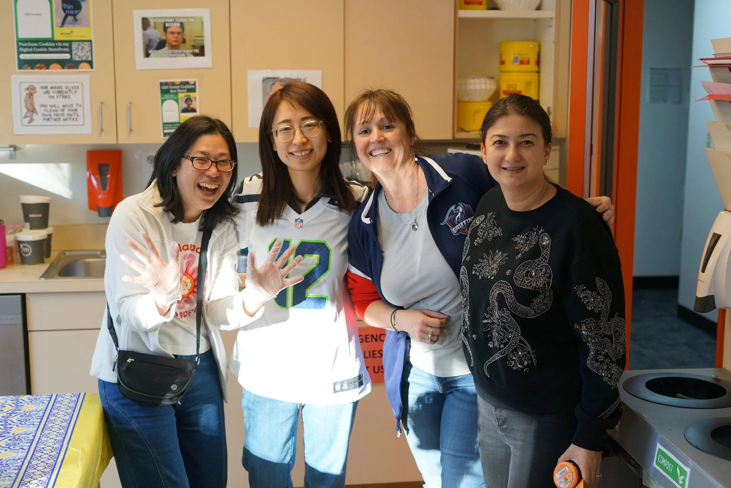Four women smiling and posing together in an indoor setting, with kitchen cabinets and posters in the background.