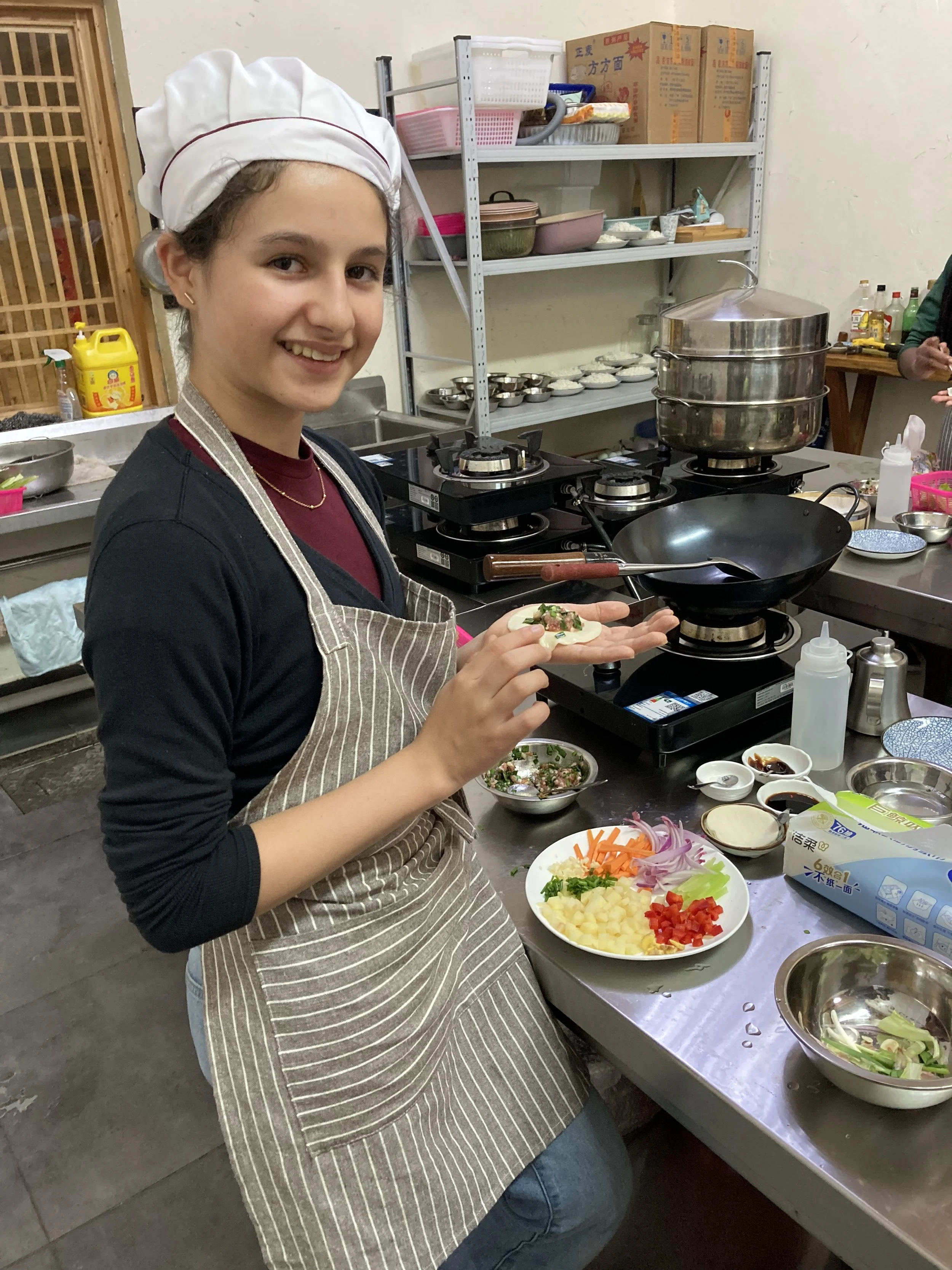 A young woman wearing a chef's hat and apron smiling while preparing food in a kitchen. She is holding a spoon, standing next to a table with chopped vegetables and various cooking utensils.