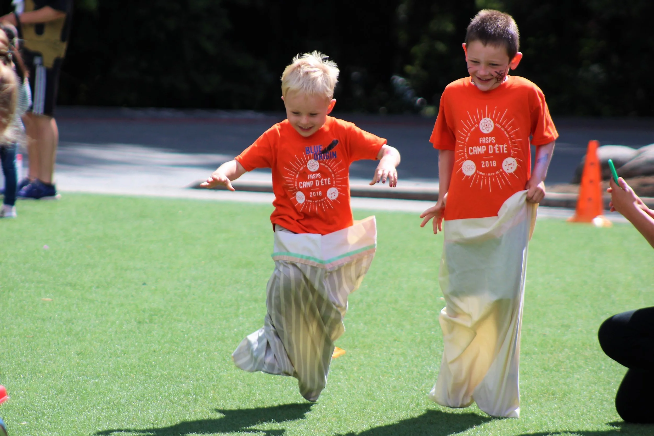 Two young boys in orange t-shirts participating in a sack race on a grass field during a summer camp event, smiling and enjoying the activity.