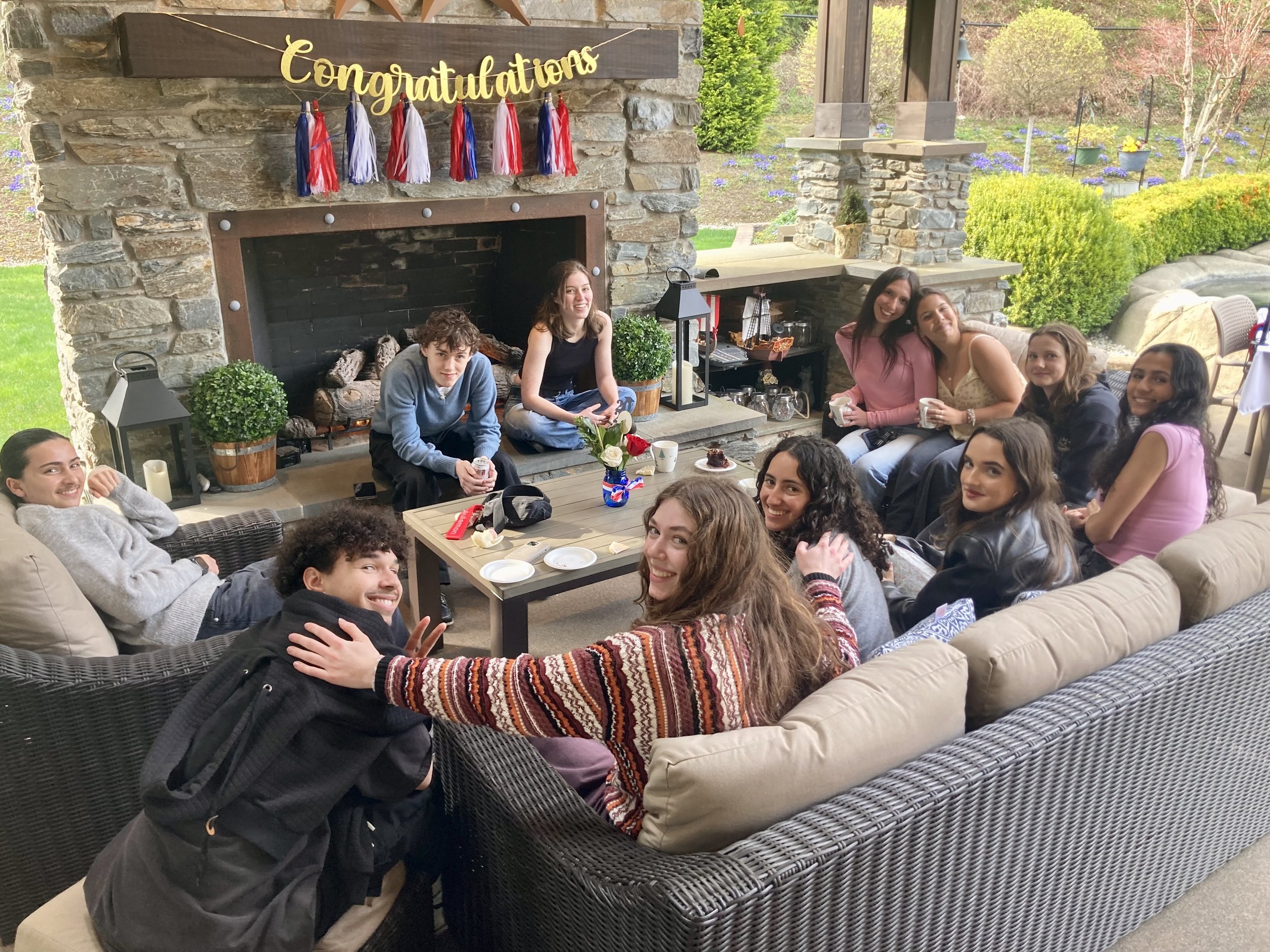 A group of young people sitting and standing around a patio with a stone fireplace, celebrating with a 'Congratulations' banner, some holding mugs, on a sunny outdoor day.