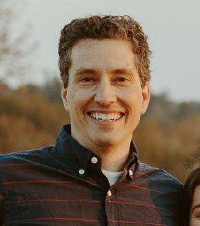 Smiling man outdoors with trees and sky in the background.