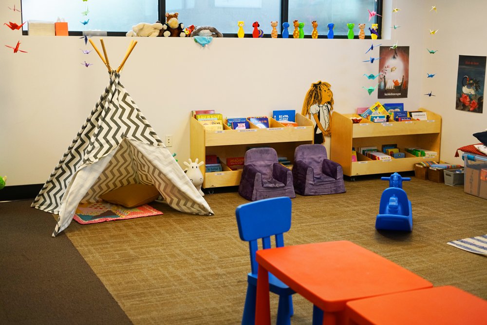 A children's play area featuring a black and white zigzag-patterned teepee tent, two purple plush armchairs, a small blue ride-on toy, a colorful table with blue and orange chairs, and a shelf filled with books against the wall. The space is decorated with wall posters, a cutout figure of a girl, and a window with a row of colorful toy giraffes and plush animals on the sill.