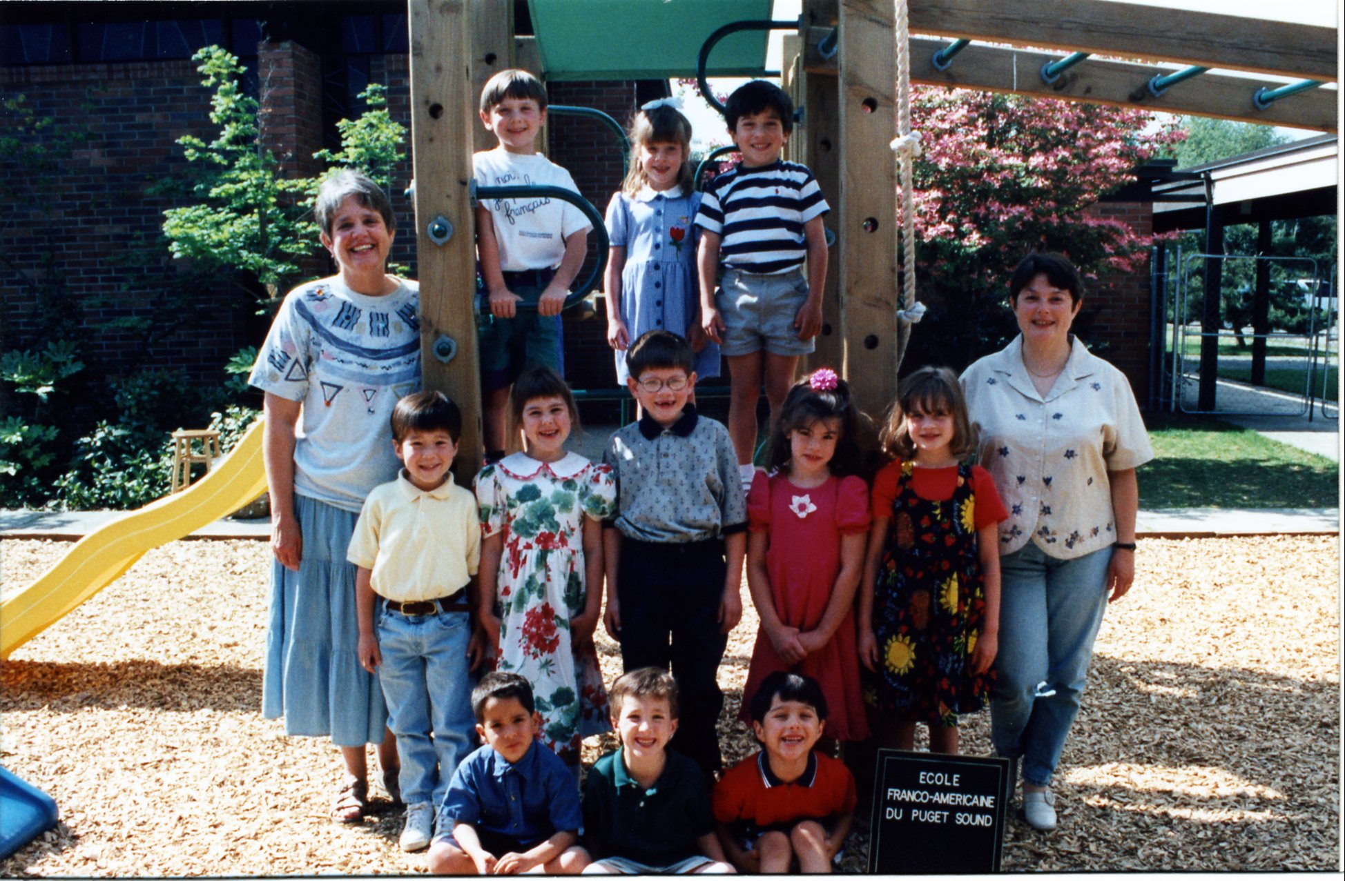 A group of children and two adults, likely teachers, posing outdoors at a playground, with a wooden play structure, slide, and a blooming pink tree in the background.