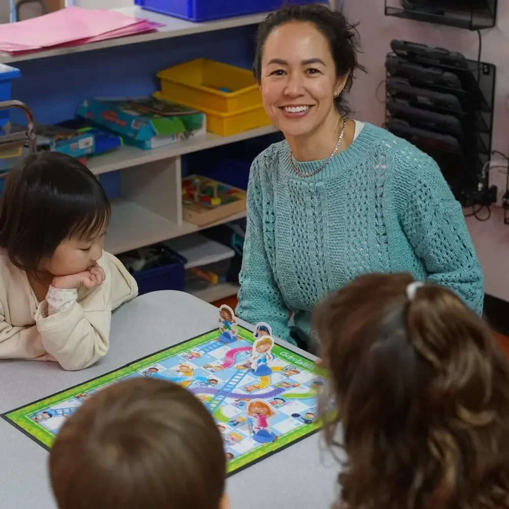A woman with short dark hair smiling at children around a table playing a board game in a classroom.