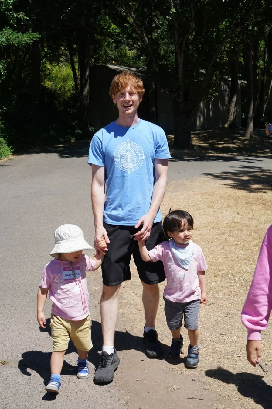A young man with red hair, wearing a blue T-shirt, is holding hands with two young children, walking outdoors on a sunny day with trees and a shed in the background.