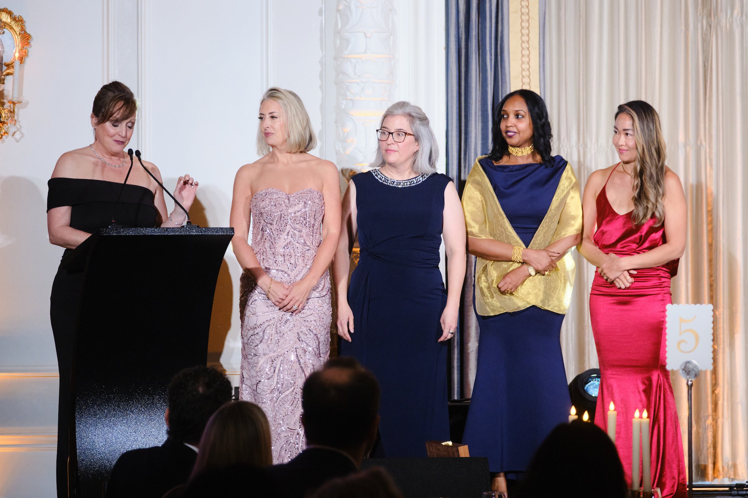 A group of women in elegant dresses standing on a stage at a formal event, with some looking at a woman speaking at a podium.