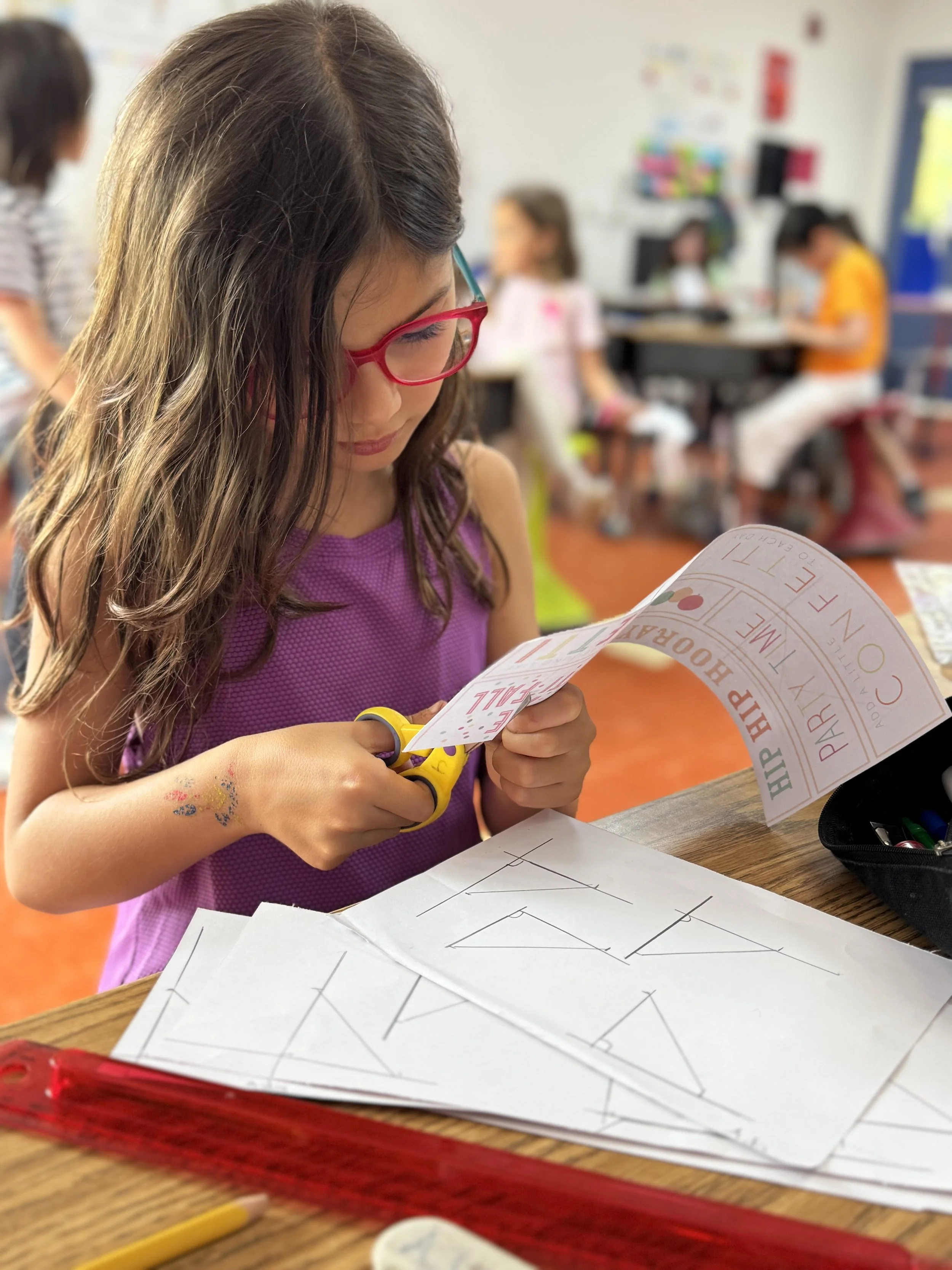 A young girl wearing glasses and a purple shirt cutting a piece of paper with yellow scissors in a classroom. There are printed worksheets and classroom supplies on the desk, with more students working in the background.
