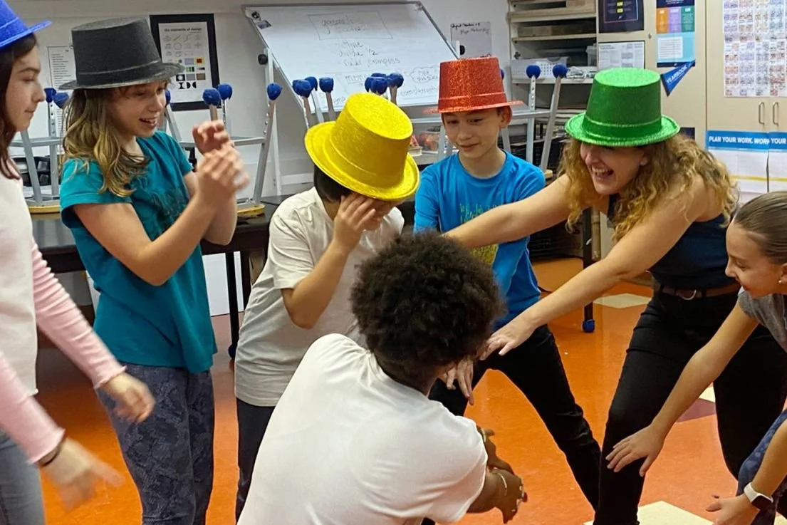 Children playing a game with a person wearing a green hat, with some kids wearing other colorful hats, in a classroom setting.
