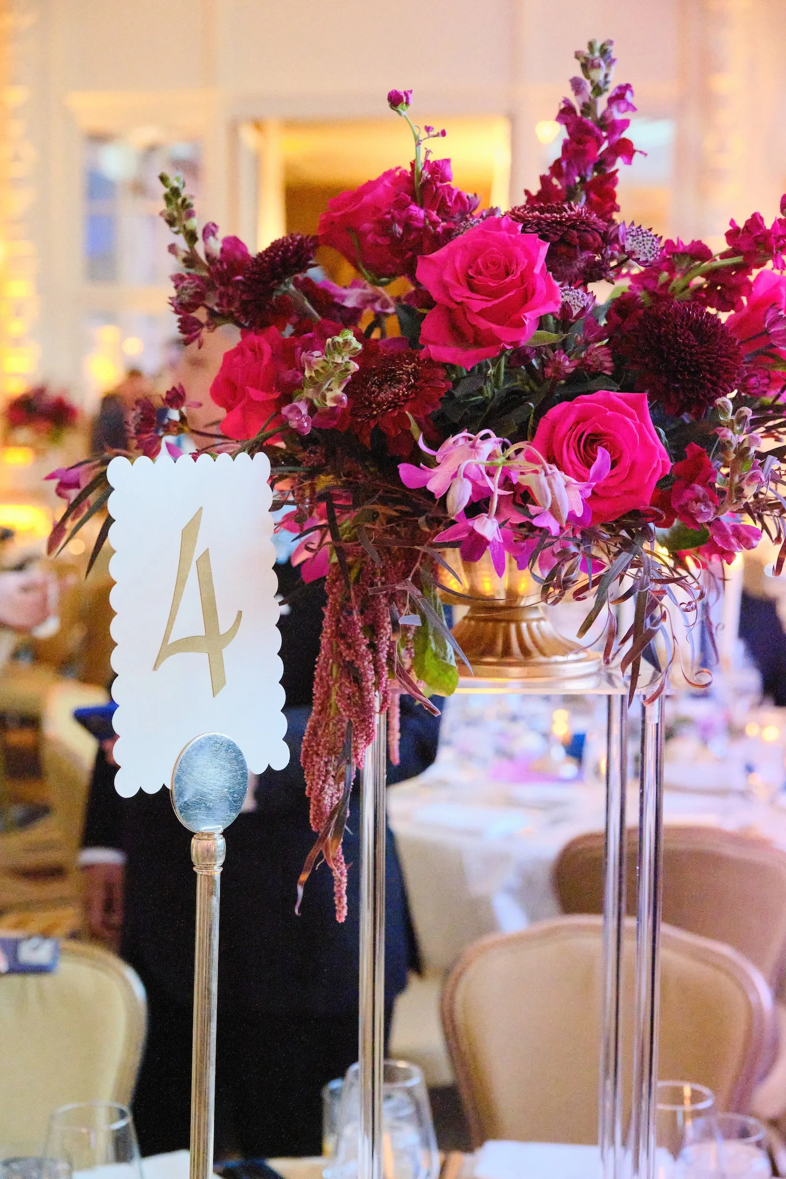 A floral centerpiece with pink and dark purple flowers on a table at a formal event, with a table number card labeled '4' attached to a stand in the foreground.