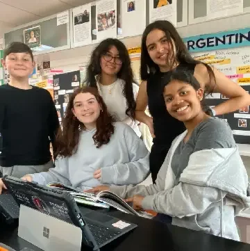 A group of children gathered around a computer in a classroom, with a teacher guiding them.