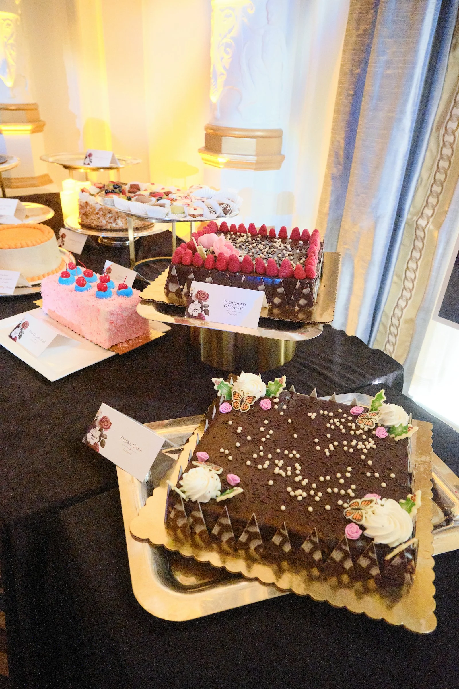 A display of decorated cakes on a table with black tablecloth, each with distinct decorations and floral small labels in the foreground.
