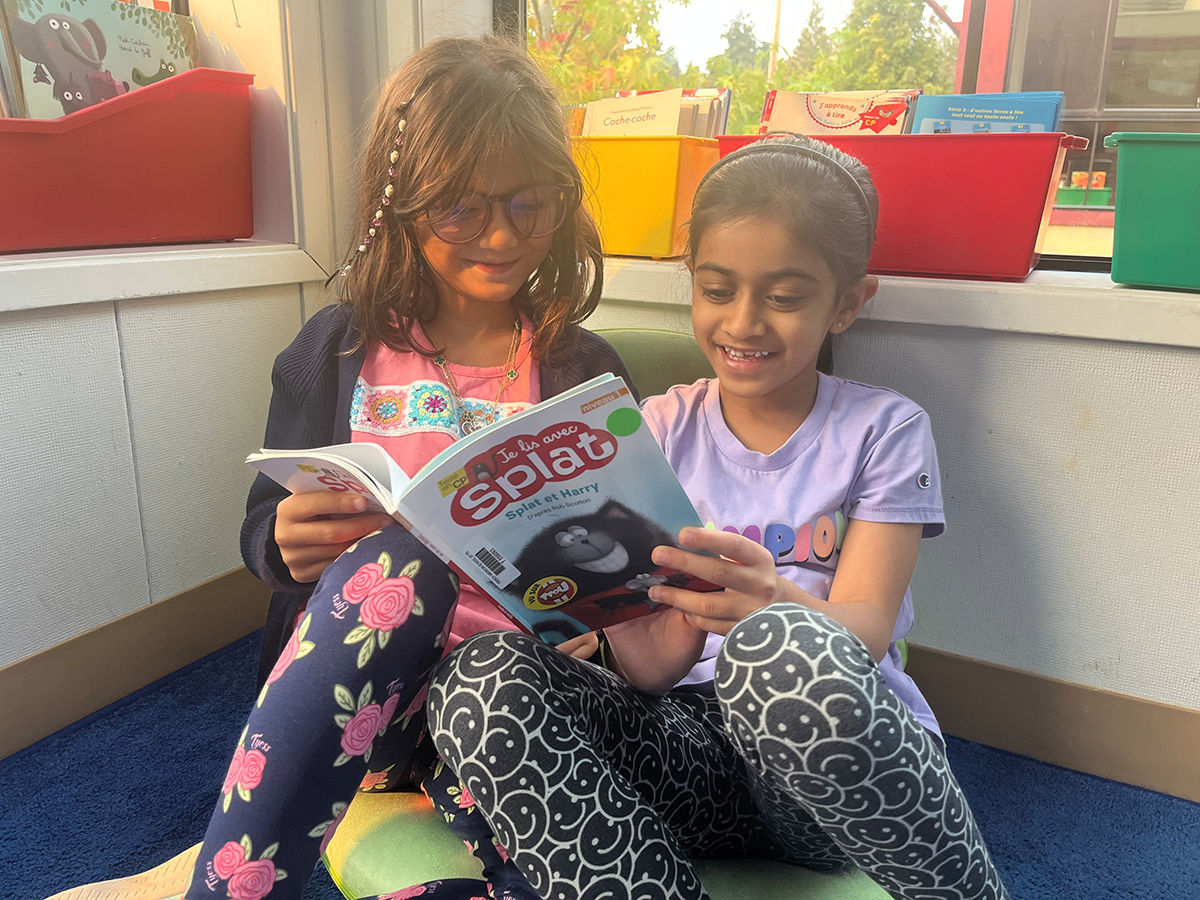 Two young girls sitting on the floor in a classroom, reading a comic book together. One girl has glasses and a beaded bracelet, the other girl has a ponytail. They are near a window with colorful storage bins and books.