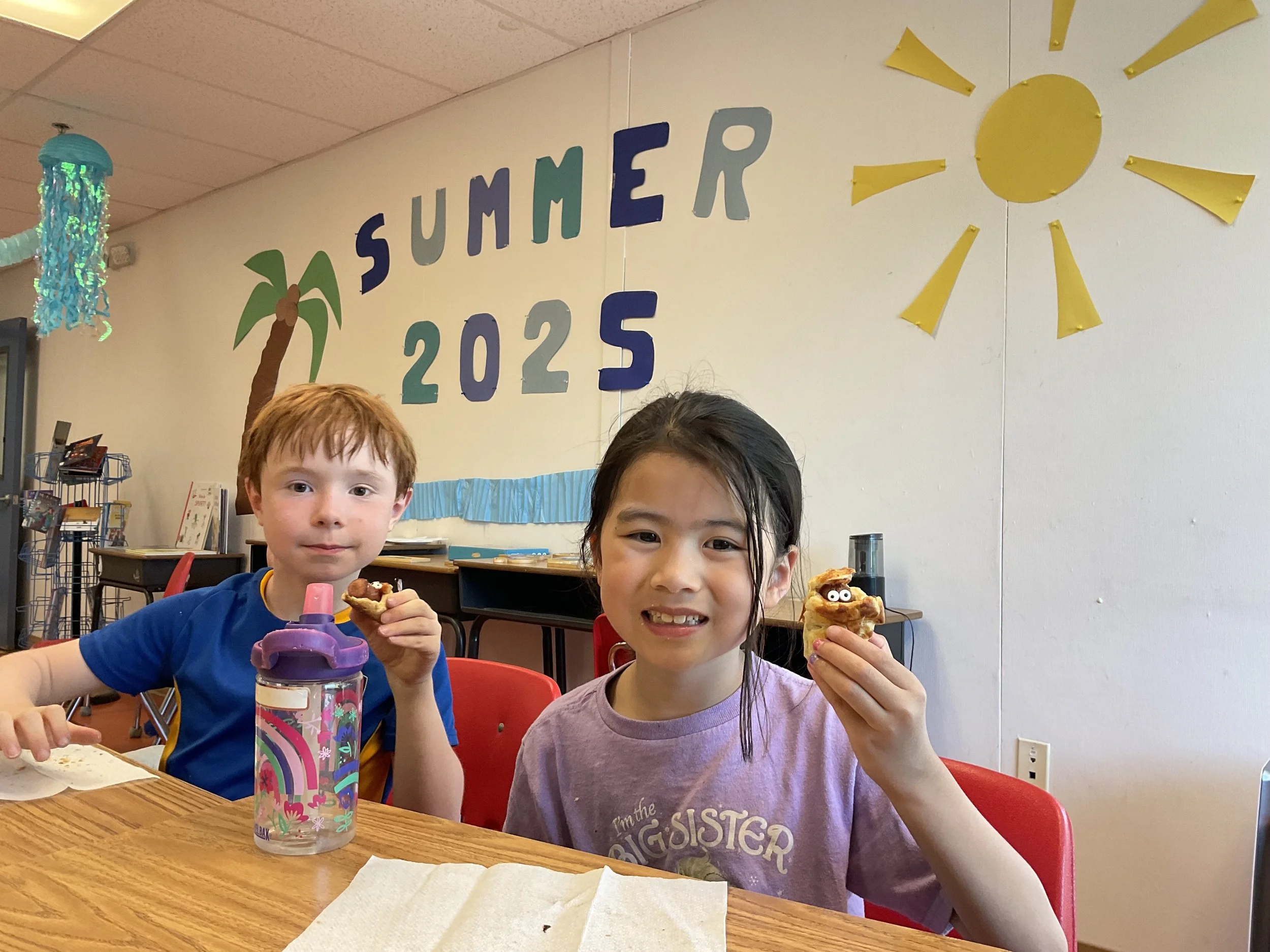 Two children sitting at a table, smiling and holding eaten cookies. A boy with red hair and a girl with dark hair. Behind them, a wall decorated with a sign reading 'SUMMER 2025' and a yellow sun, indicating a summer celebration or event.