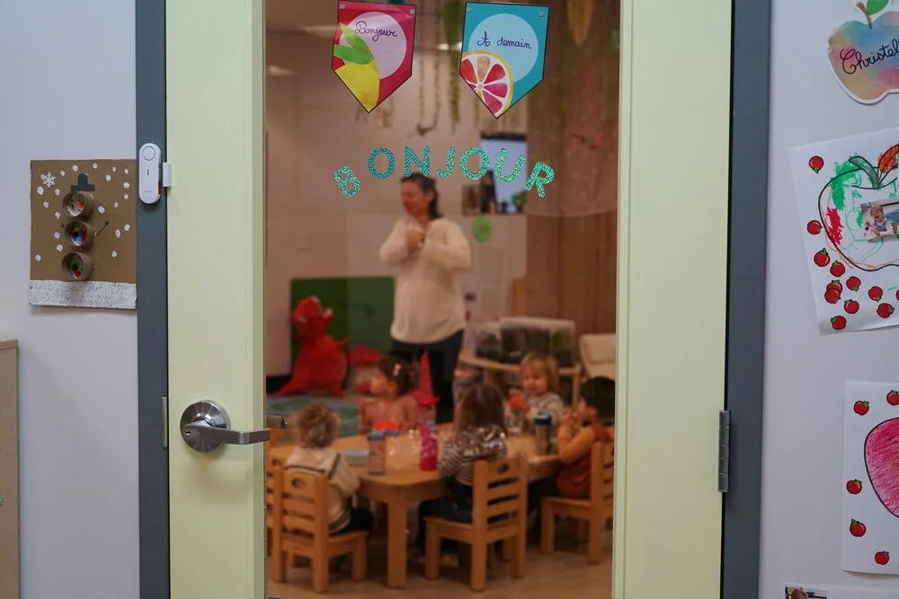 A classroom door with holiday decorations and a 'Bonjour' sign, showing children sitting around a small table inside, with a teacher standing in the background. The room is decorated with colorful banners and drawings.