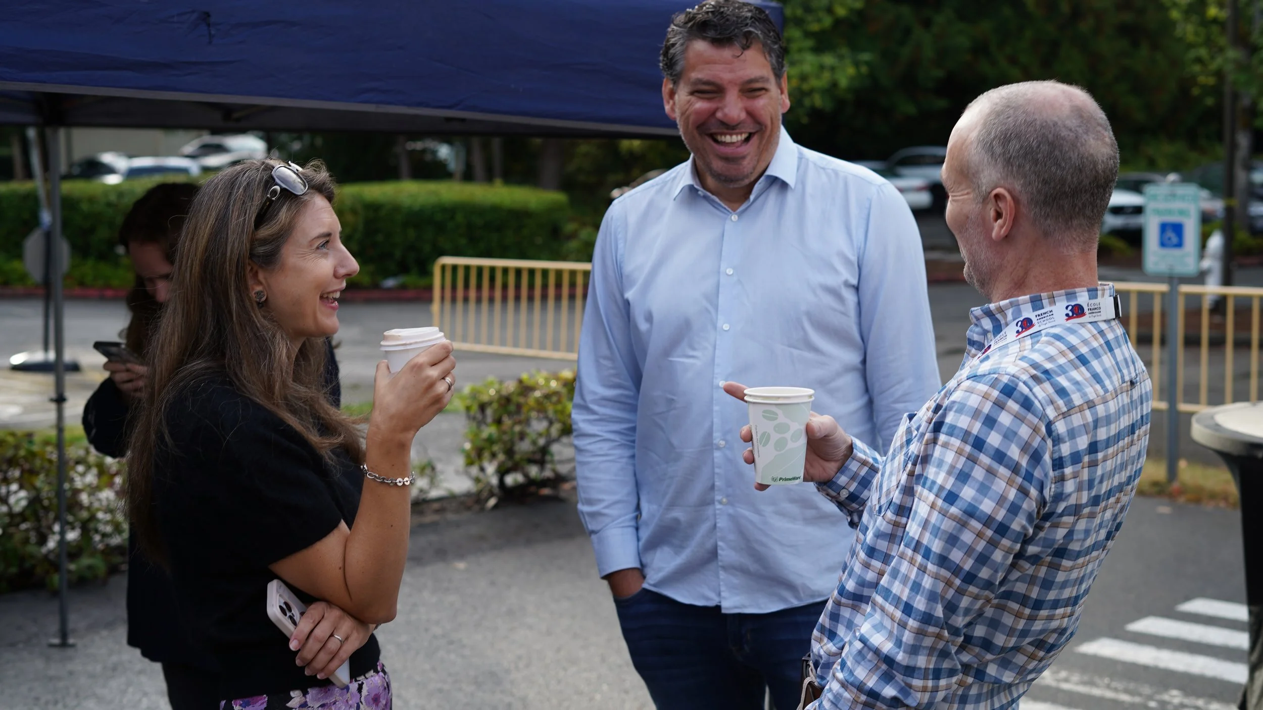 Three people smiling and conversing outdoors, two holding disposable coffee cups, a woman with long brown hair and a man with a shaved head, dressed casually, standing under a blue canopy with parked cars and greenery in the background.